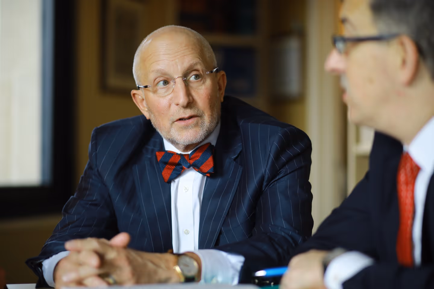 Two elderly men in suits having a serious conversation, one wearing a red and navy striped bow tie and glasses.