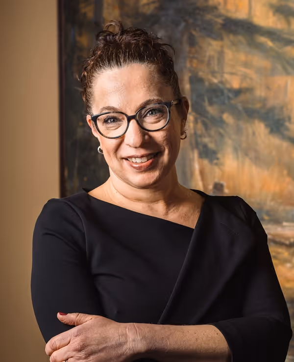 Smiling woman with glasses and hair tied up, wearing a black top, standing with arms crossed.