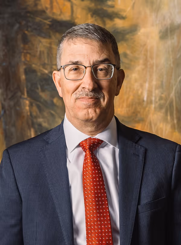 Middle-aged man with glasses, gray hair, and a mustache wearing a dark suit, white shirt, and red patterned tie against a blurred warm-toned background.