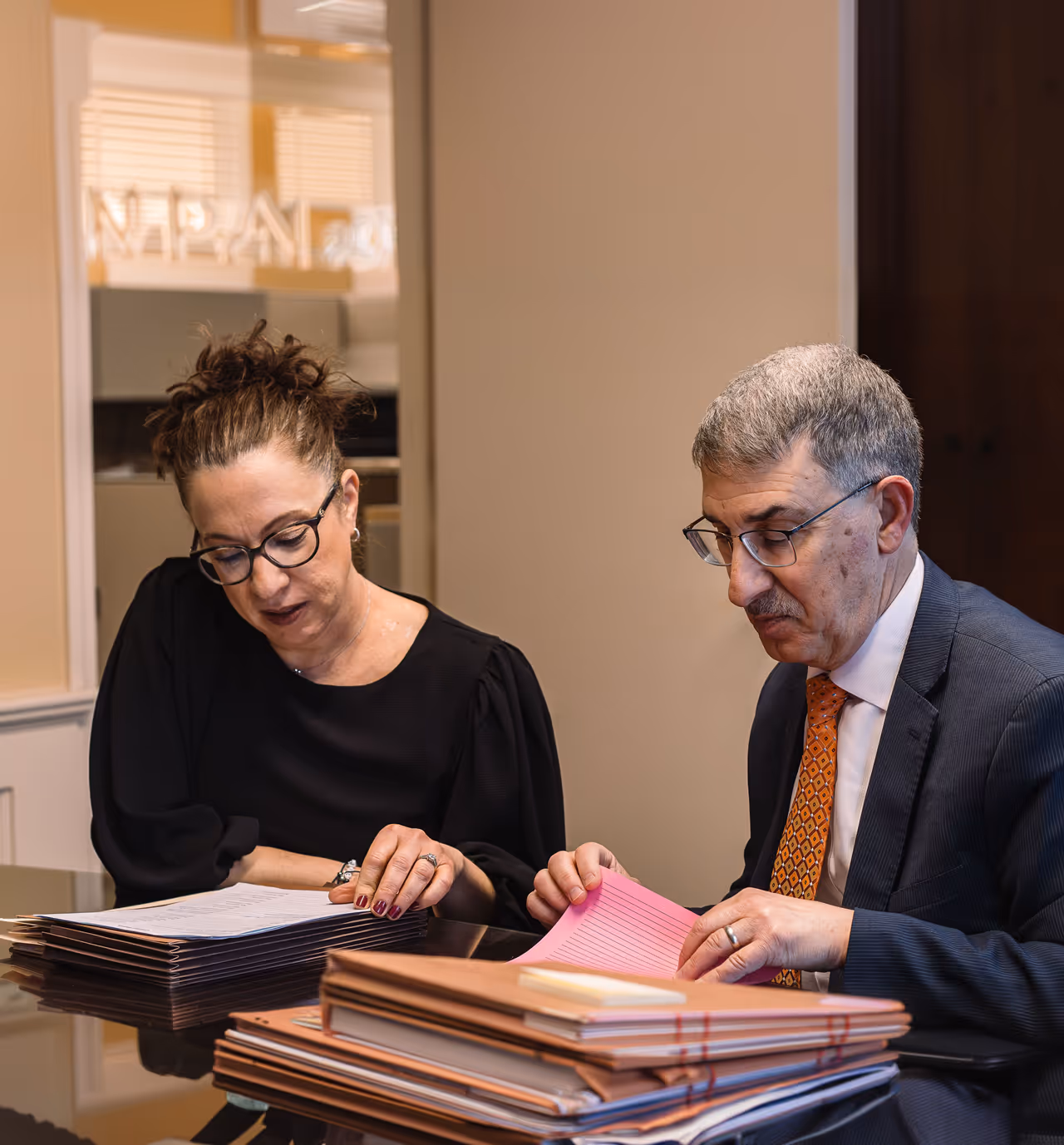 Two professionals sitting at a desk, reviewing stacks of documents, with the woman on the left wearing glasses and a black top, and the man on the right in a suit and orange patterned tie.