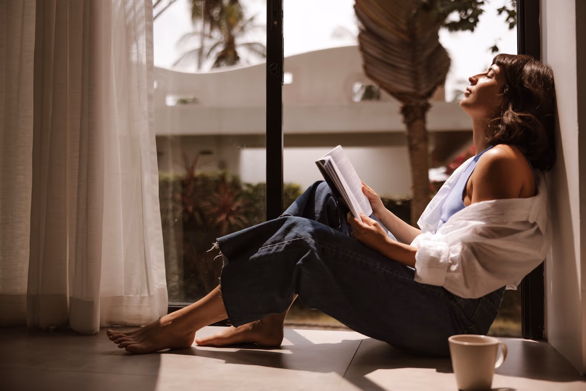 A woman sitting comfortably on a sunlit floor by a large window, leaning against a wall and reading a book with a coffee cup nearby.