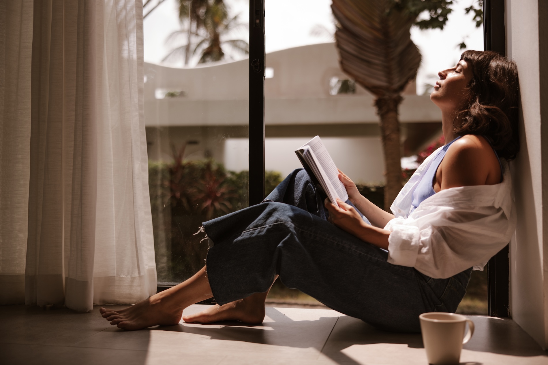 A woman sitting comfortably on a sunlit floor by a large window, leaning against a wall and reading a book with a coffee cup nearby.