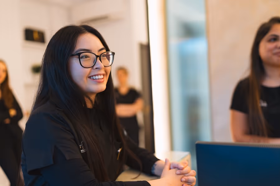 Smiling woman with long black hair and glasses sitting at a desk in an office environment.