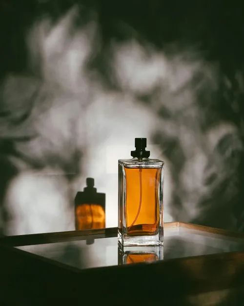 Rectangular glass perfume bottle with amber liquid on a reflective surface casting a shadow on a blurred background.