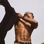 Shirtless man wearing sunglasses and a watch, adjusting a black jacket over his shoulder against a clear sky.