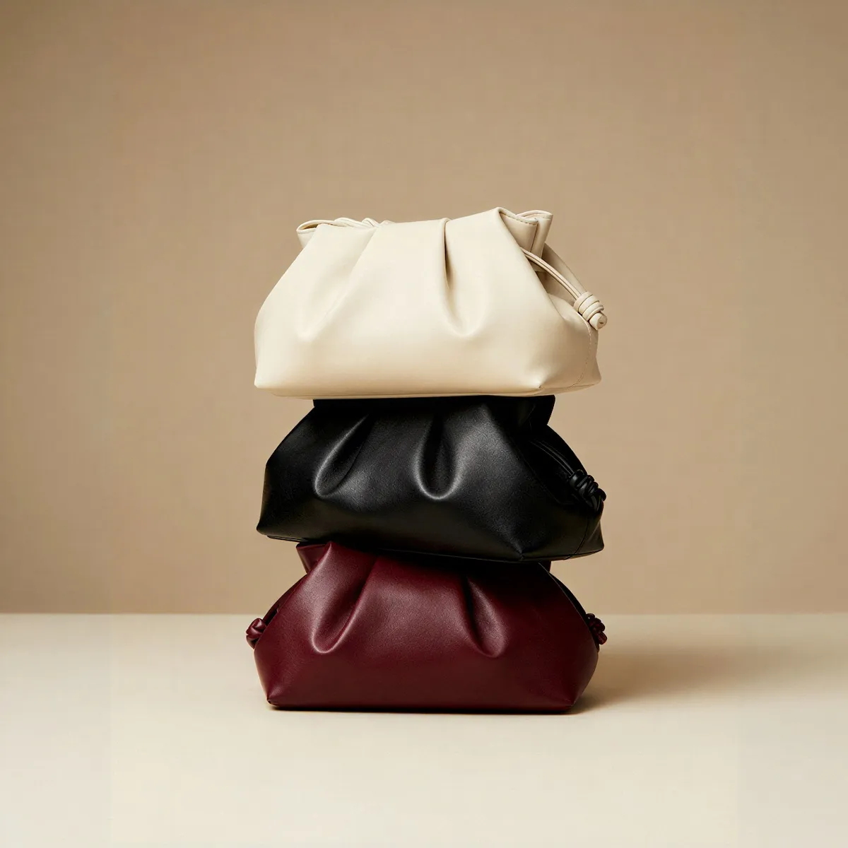 Stack of three soft leather pouches in cream, black, and burgundy colors on a beige surface and background.