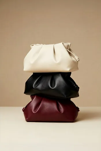 Stack of three soft leather clutch bags in beige, black, and burgundy on a beige background.