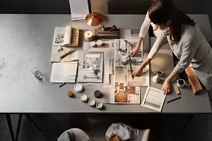 A designer working at a desk with architectural plans, sketches, color samples, and a laptop.