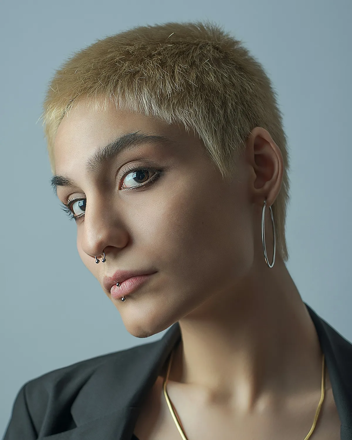 Close-up portrait of a person with short blond hair, facial piercings, hoop earring, and a black blazer against a plain background.