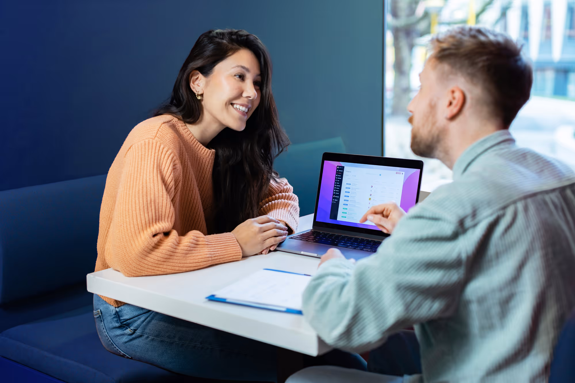 Zwei Personen sitzen an einem Tisch, eine zeigt etwas auf einem Laptopbildschirm, beide lächeln.