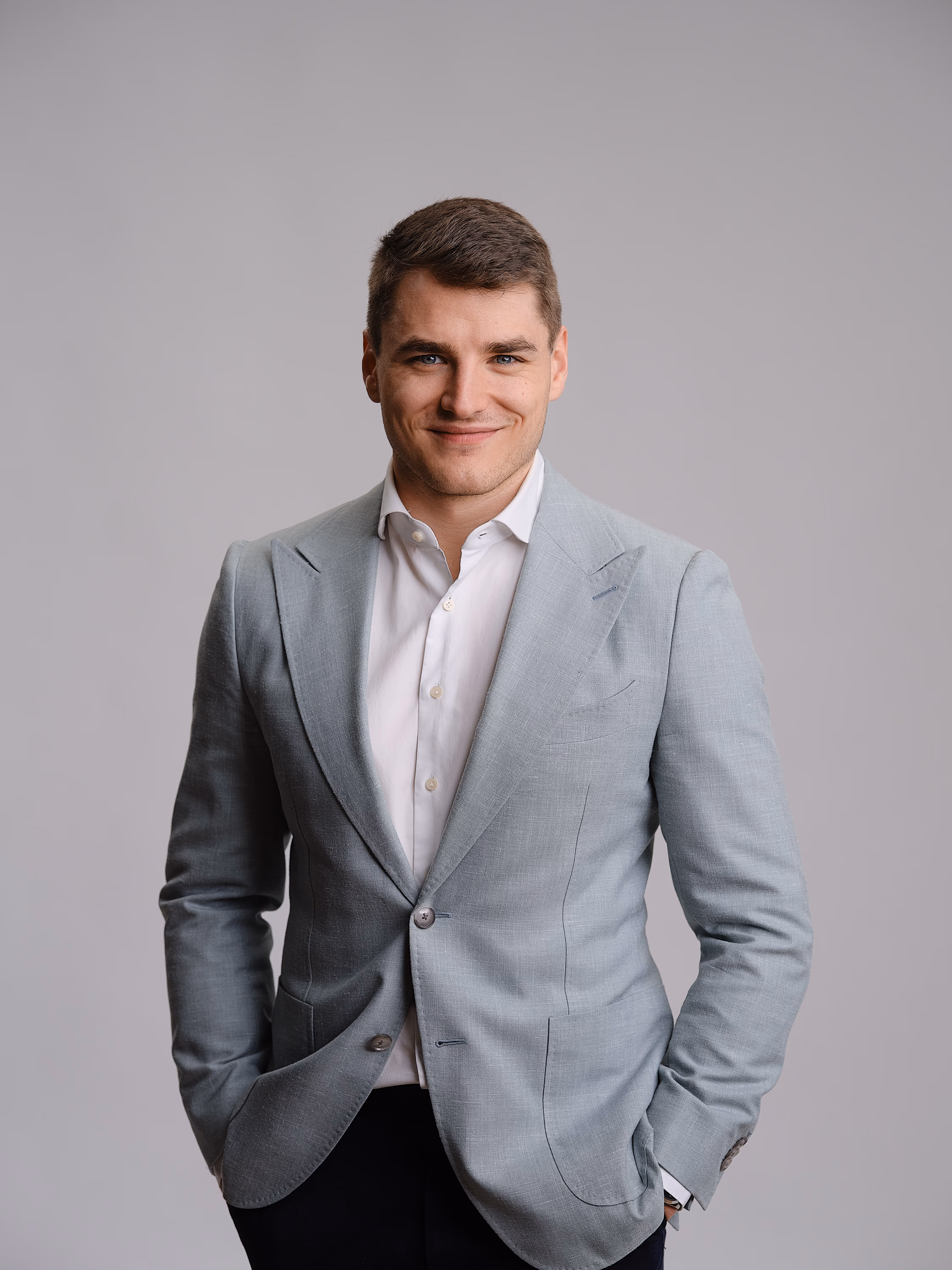 Junger Mann mit hellgrauem Sakko, weißem Hemd und dunkler Hose vor grauem Hintergrund, Hände in den Taschen.
