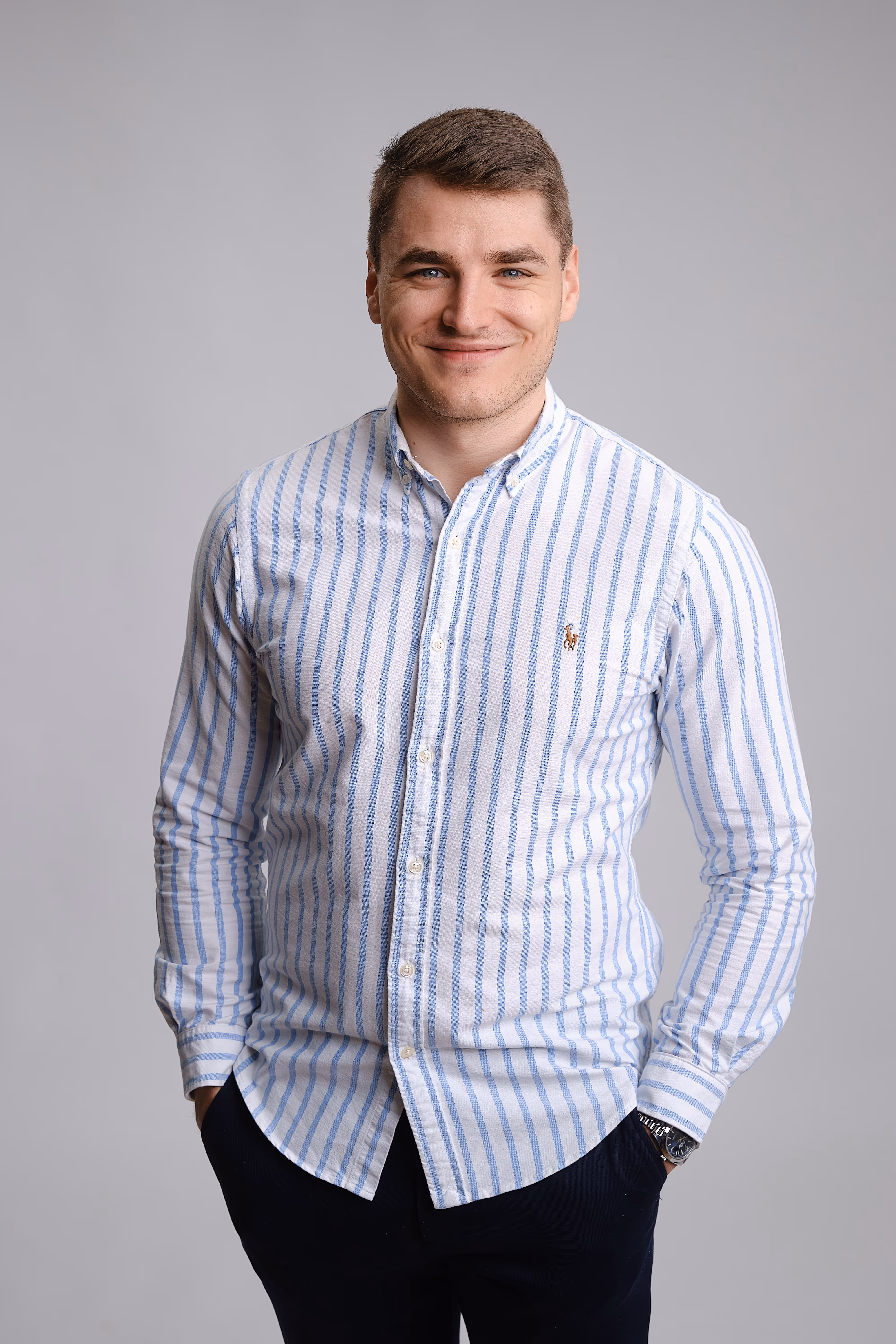 Lächelnder Mann mit kurz geschnittenem braunem Haar trägt ein weißes Hemd mit blauen Streifen und schwarze Hose vor einem grauen Hintergrund.