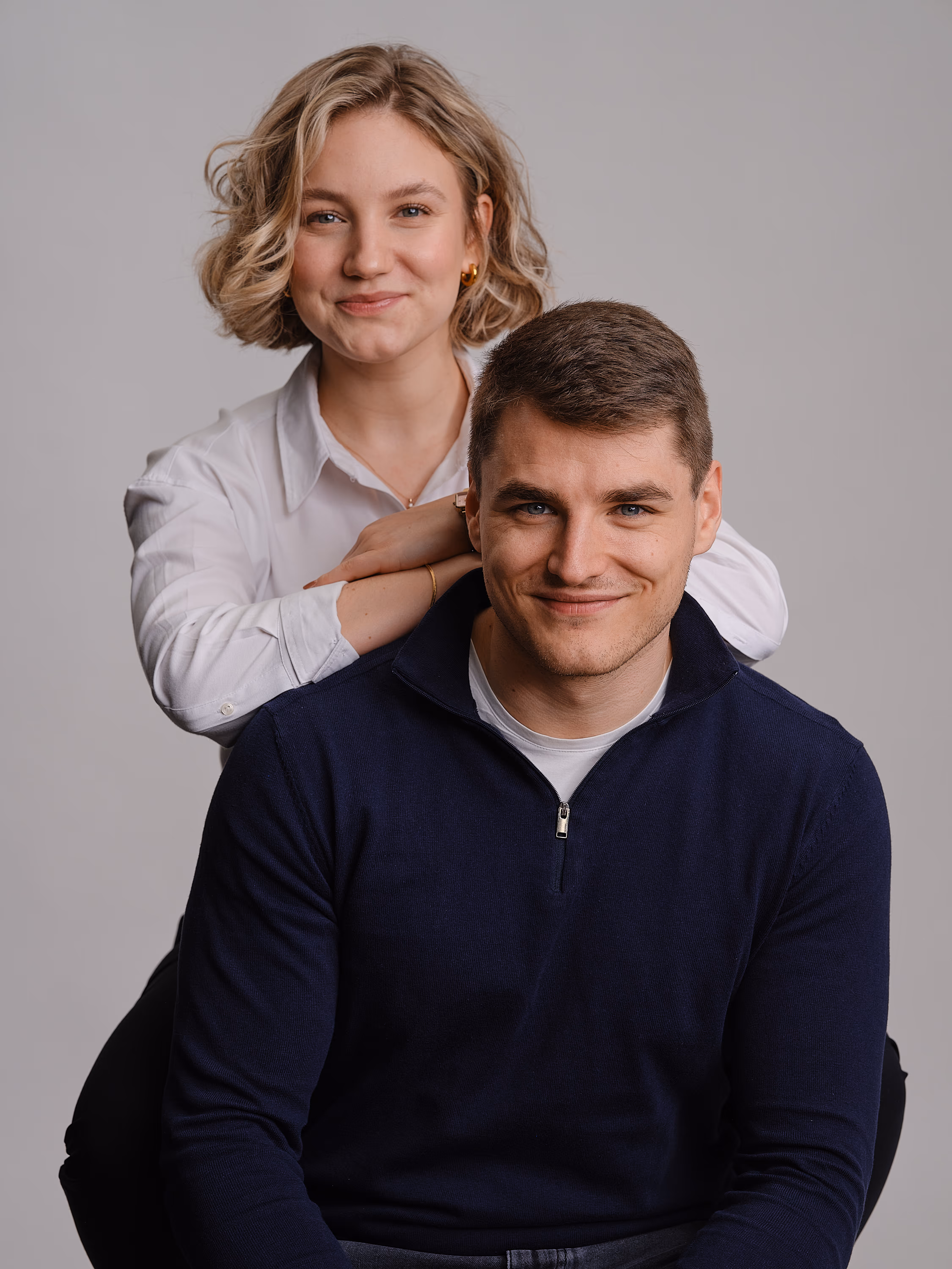 Porträt eines lächelnden jungen Mannes in einem dunkelblauen Pullover mit einer lächelnden Frau mit blonden kurzen Haaren, die hinter ihm steht und ihre Arme auf seine Schultern legt.