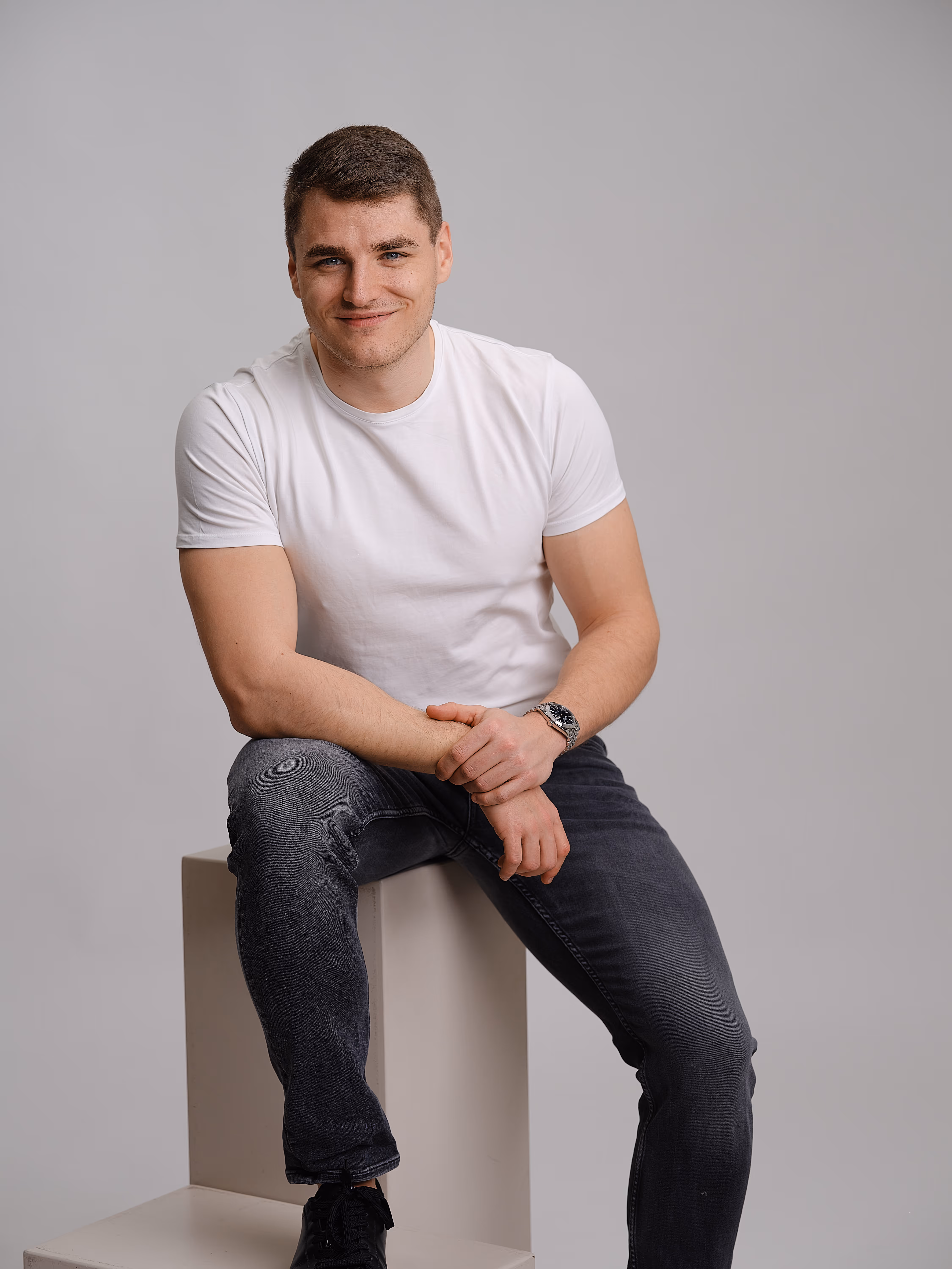 Junger Mann mit weißem T-Shirt, dunklen Jeans und Armbanduhr sitzt lächelnd auf einem Hocker vor grauem Hintergrund.