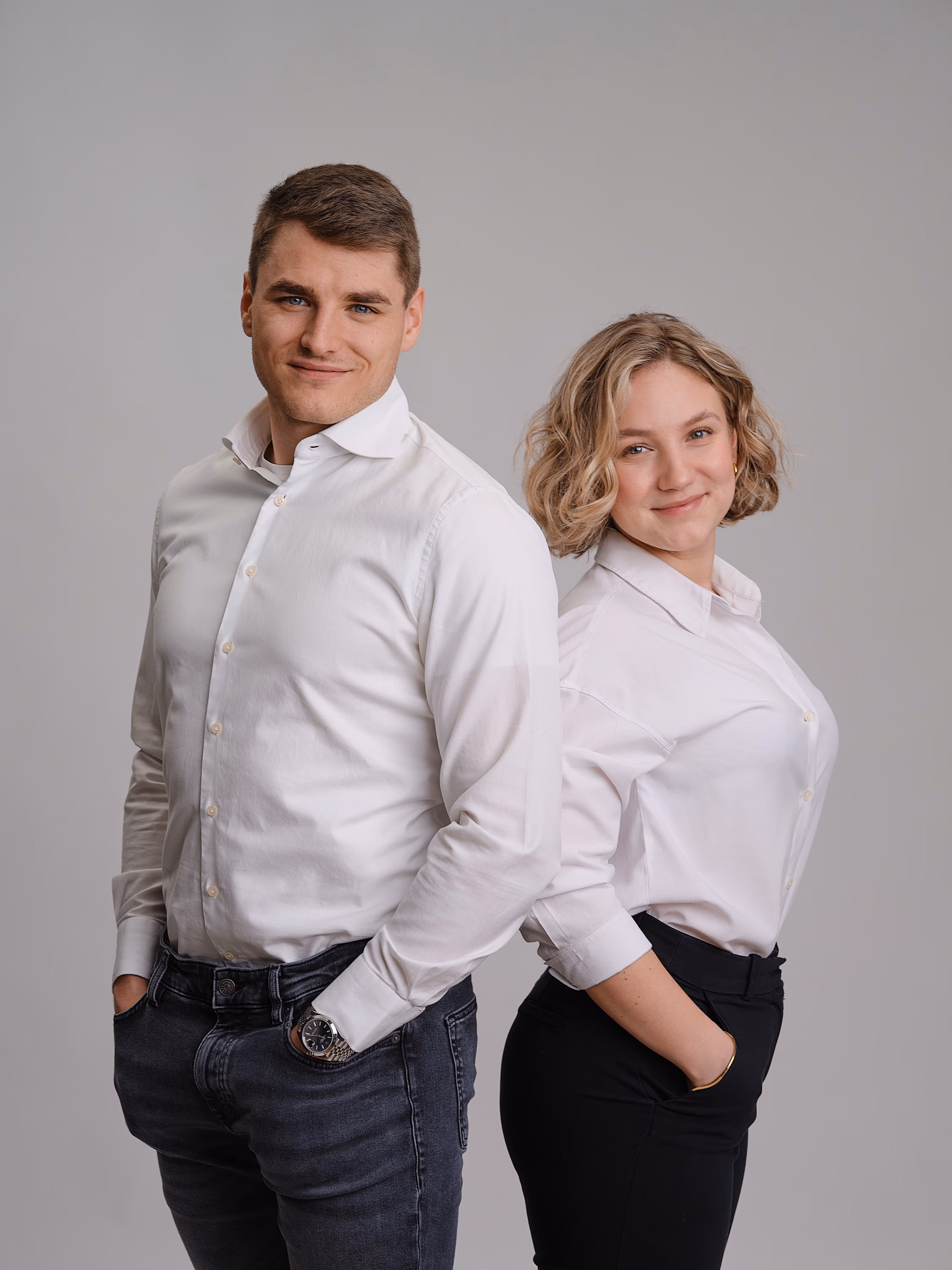 Junger Mann und junge Frau stehen Rücken an Rücken, beide tragen weiße Hemden und lächeln.