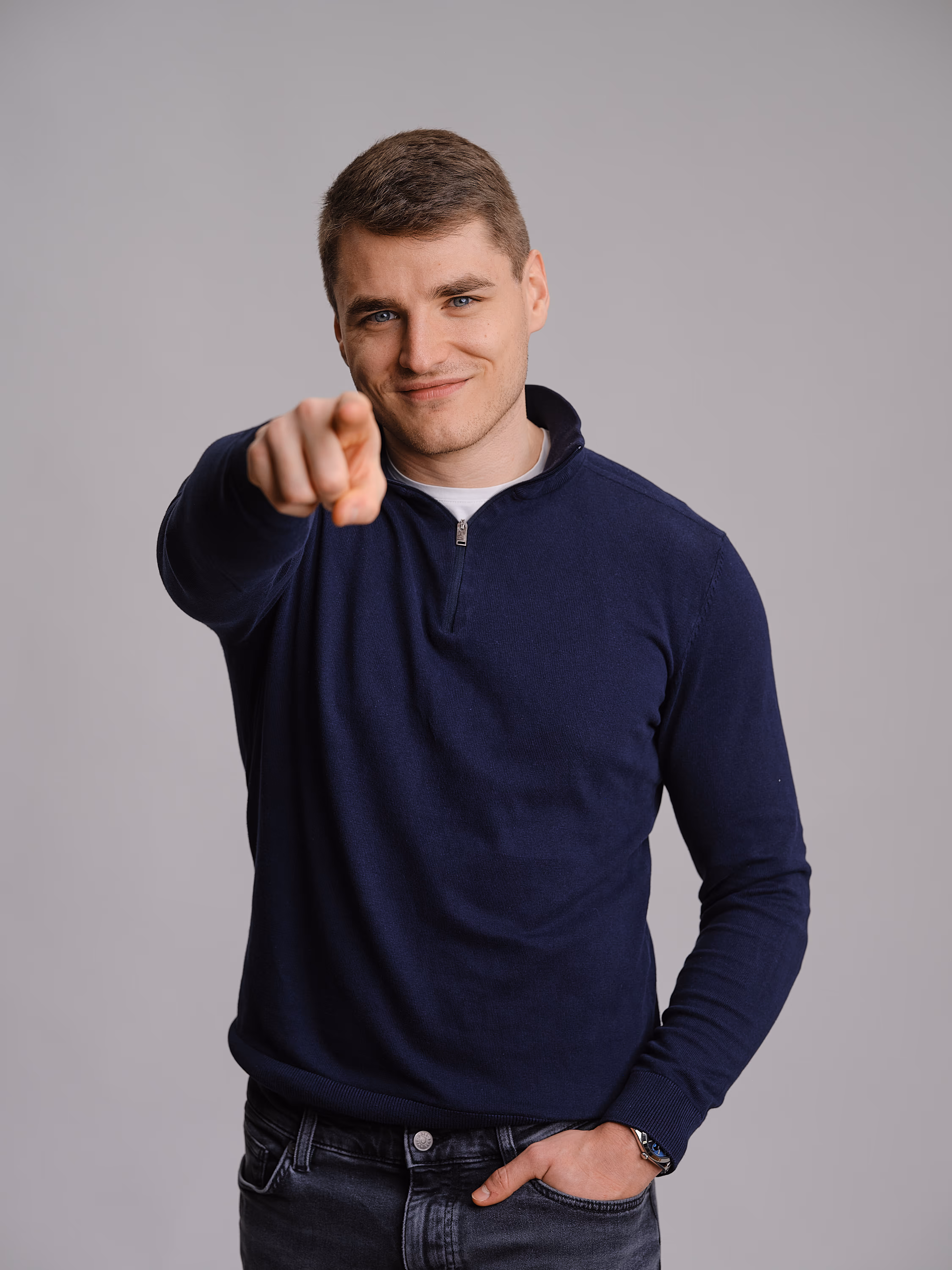Junger Mann mit blauem Pullover zeigt direkt auf den Betrachter und lächelt.