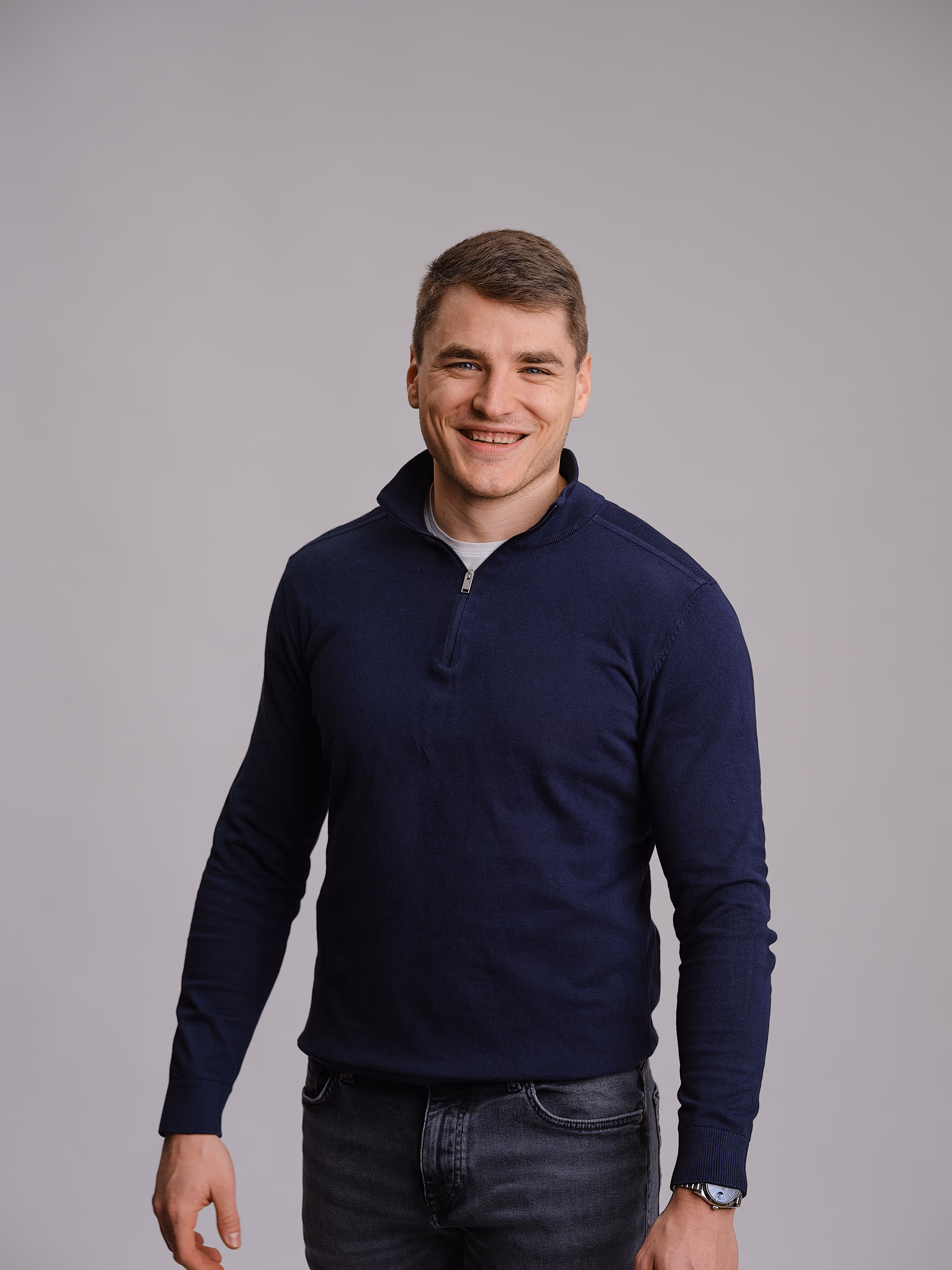 Junger Mann mit kurzem braunem Haar, der ein dunkles Sweatshirt und eine Armbanduhr trägt und lächelt.