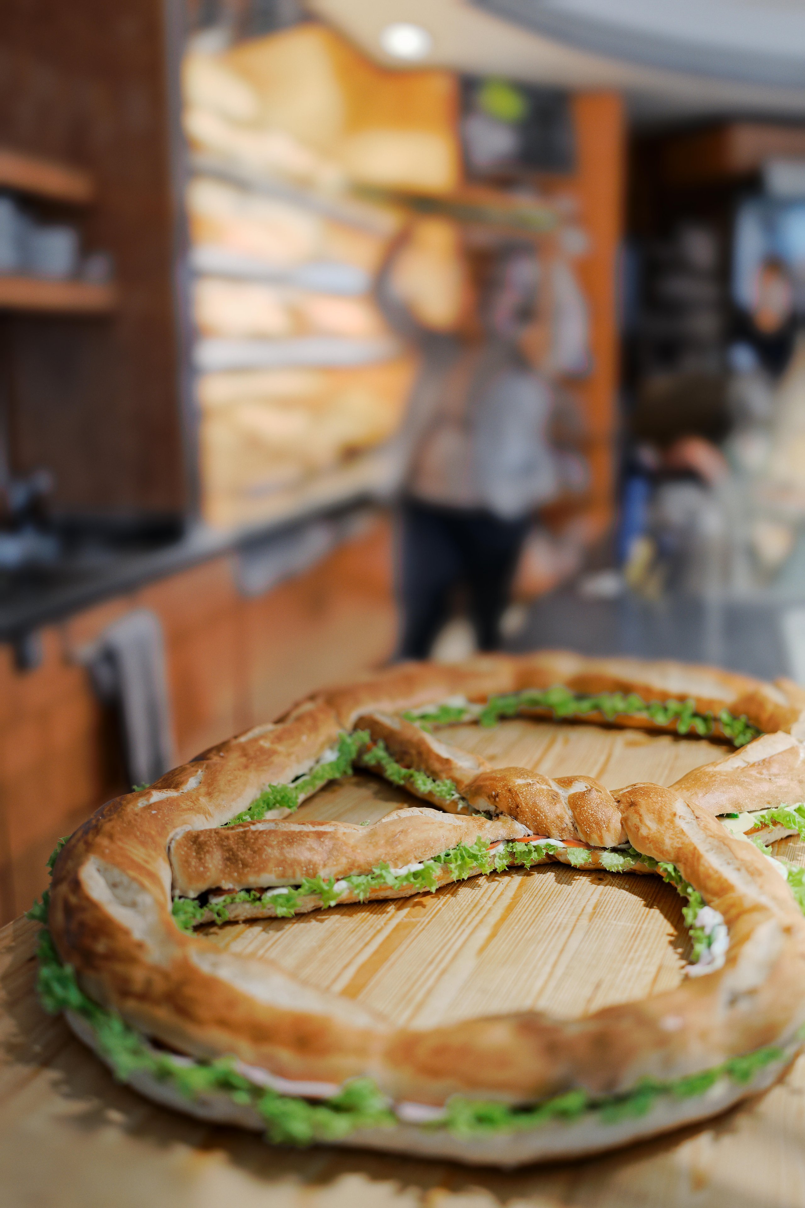 Large sandwich shaped like a peace symbol with lettuce on a wooden surface in a bakery setting.