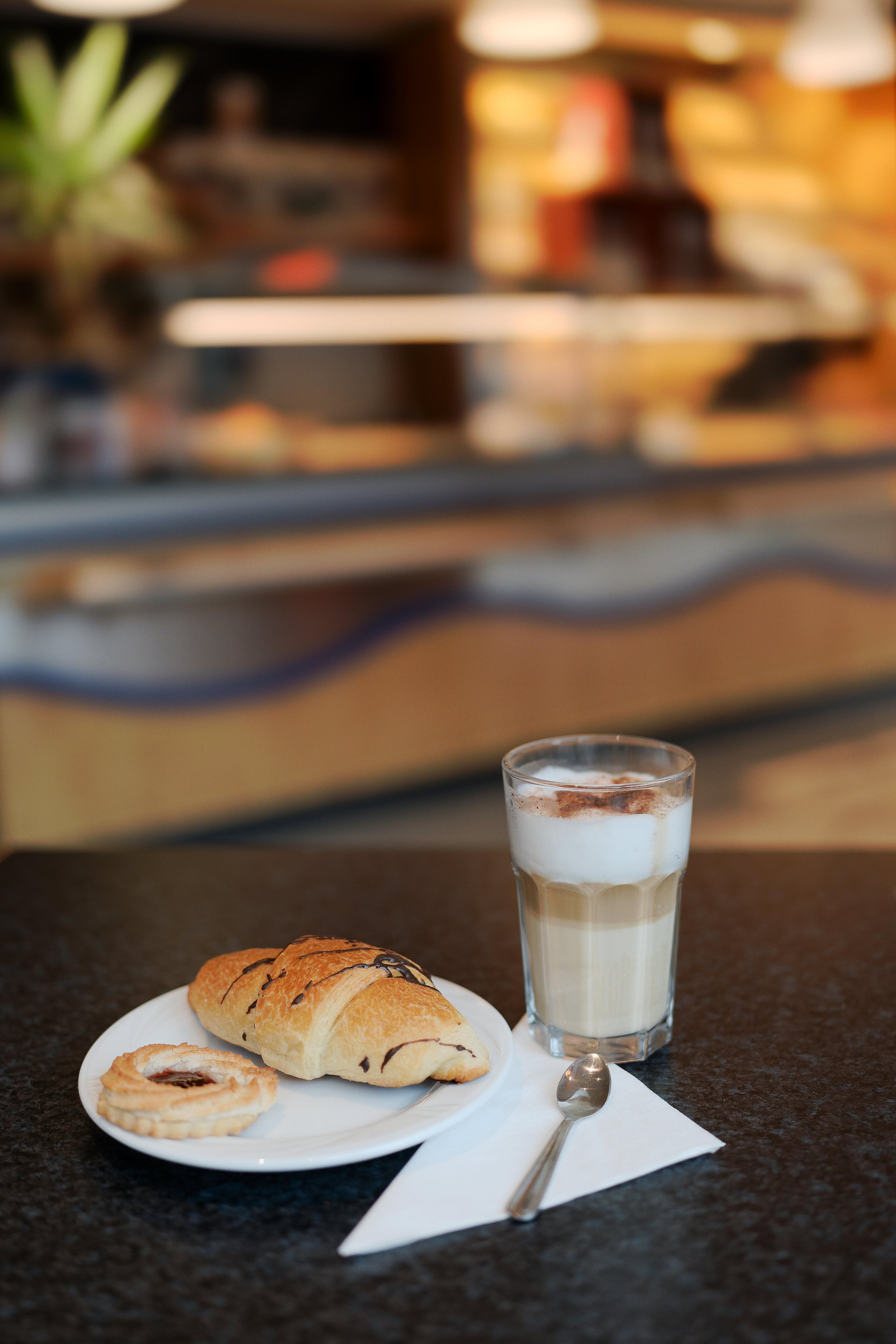 Glass of cappuccino with foam and cinnamon on a black table next to a plate with a chocolate-drizzled croissant and a cookie, with a spoon on a white napkin.