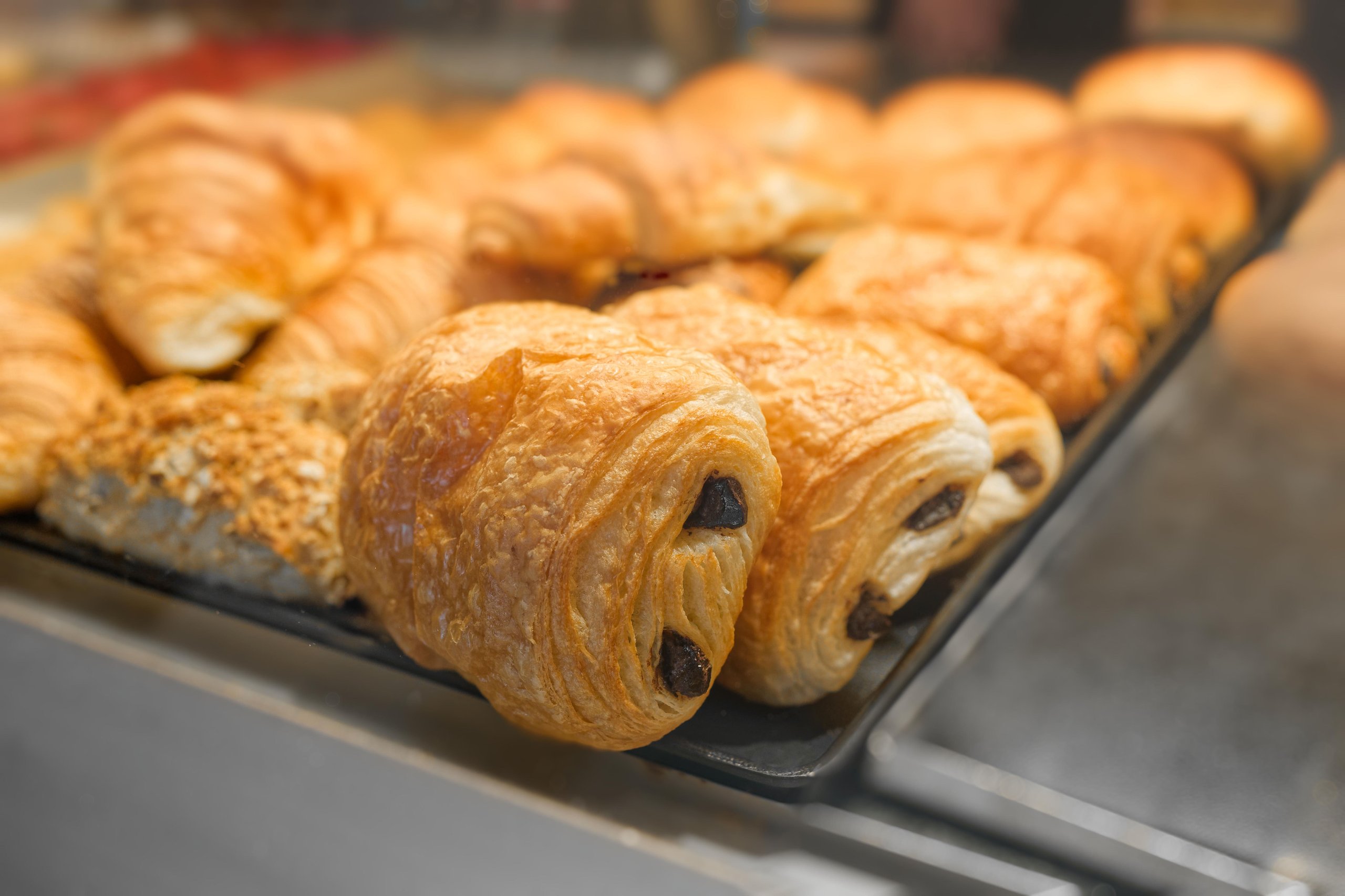 Tray of freshly baked golden brown chocolate croissants and assorted pastries in a bakery display.