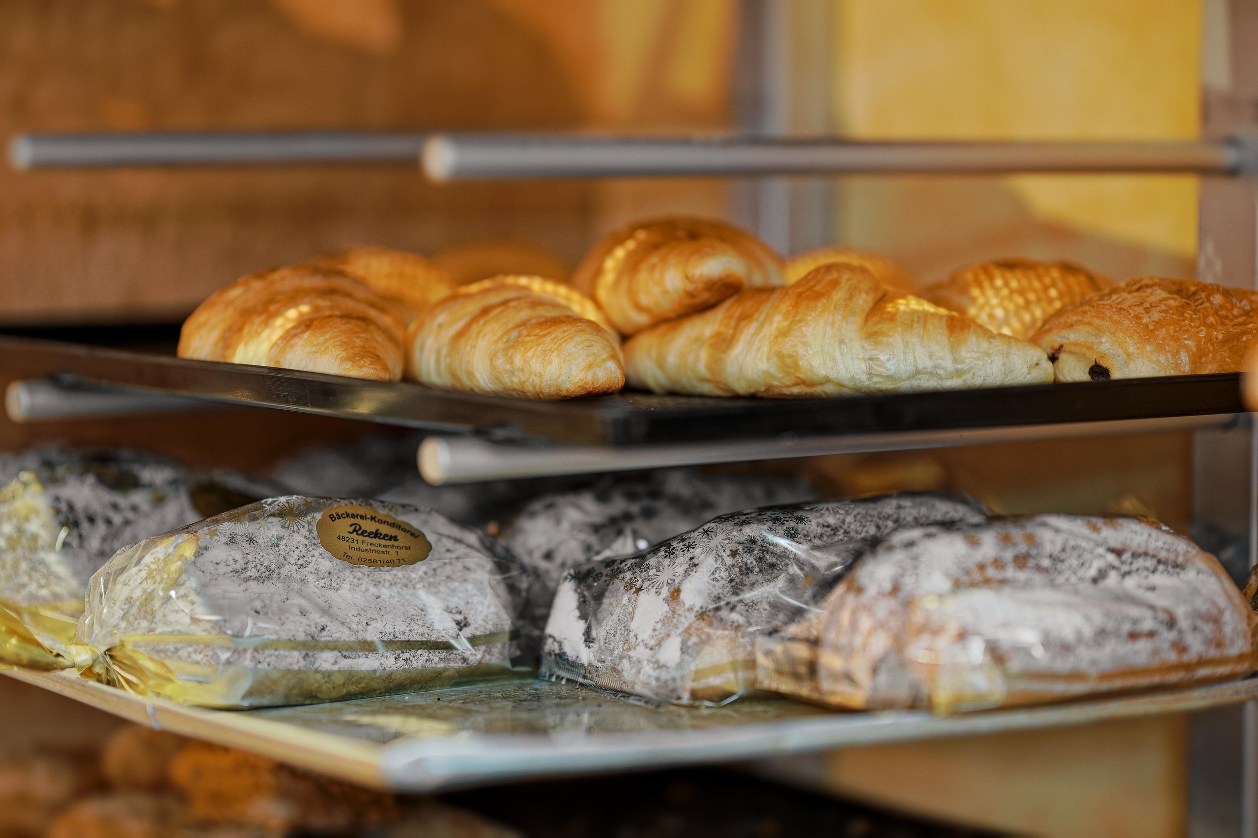 Bakery shelf displaying golden croissants on the top tray and powdered sugar-covered pastries wrapped in clear plastic on the middle tray.