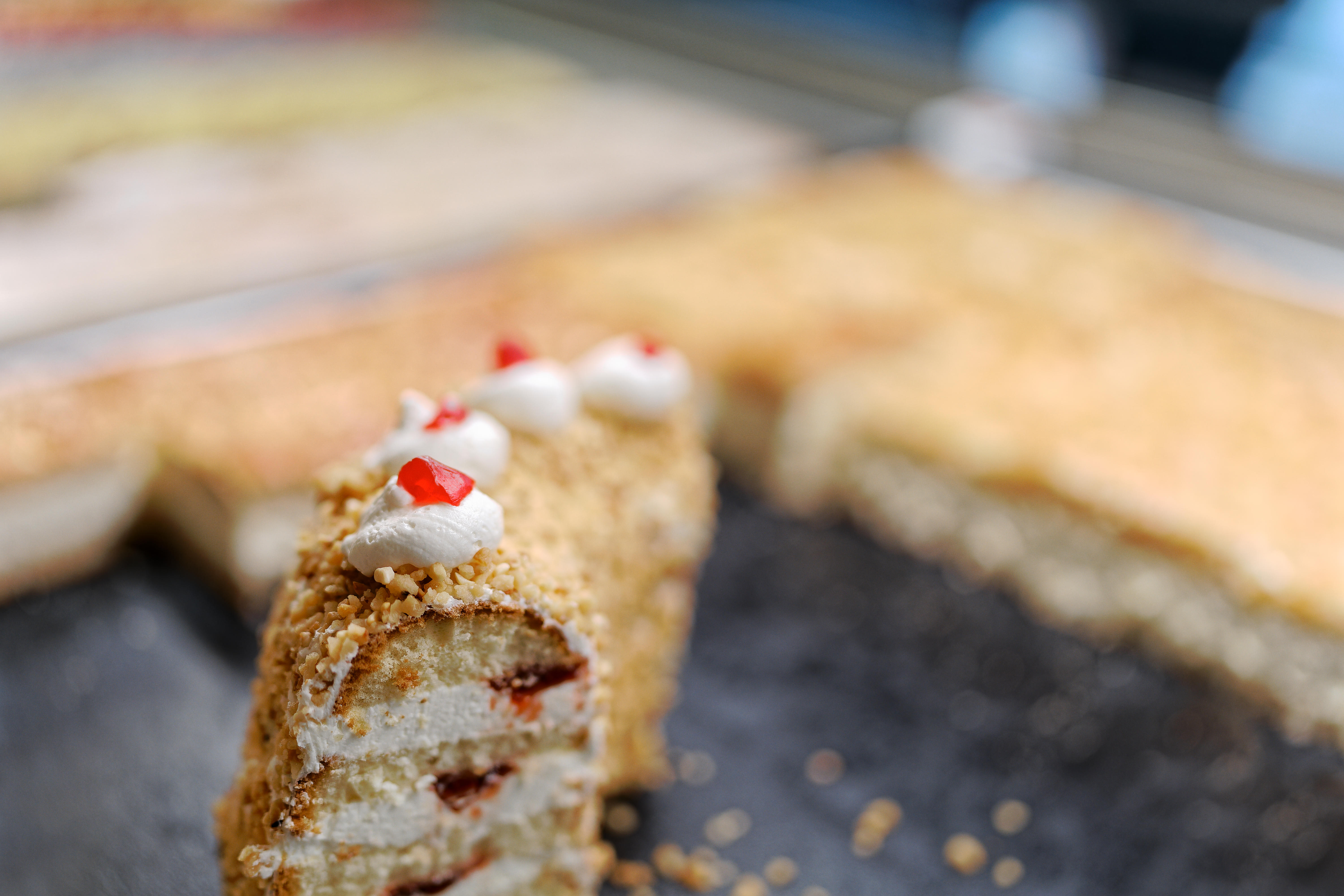 Slice of layered cake with cream, nuts, and red jelly toppings on a dark surface.
