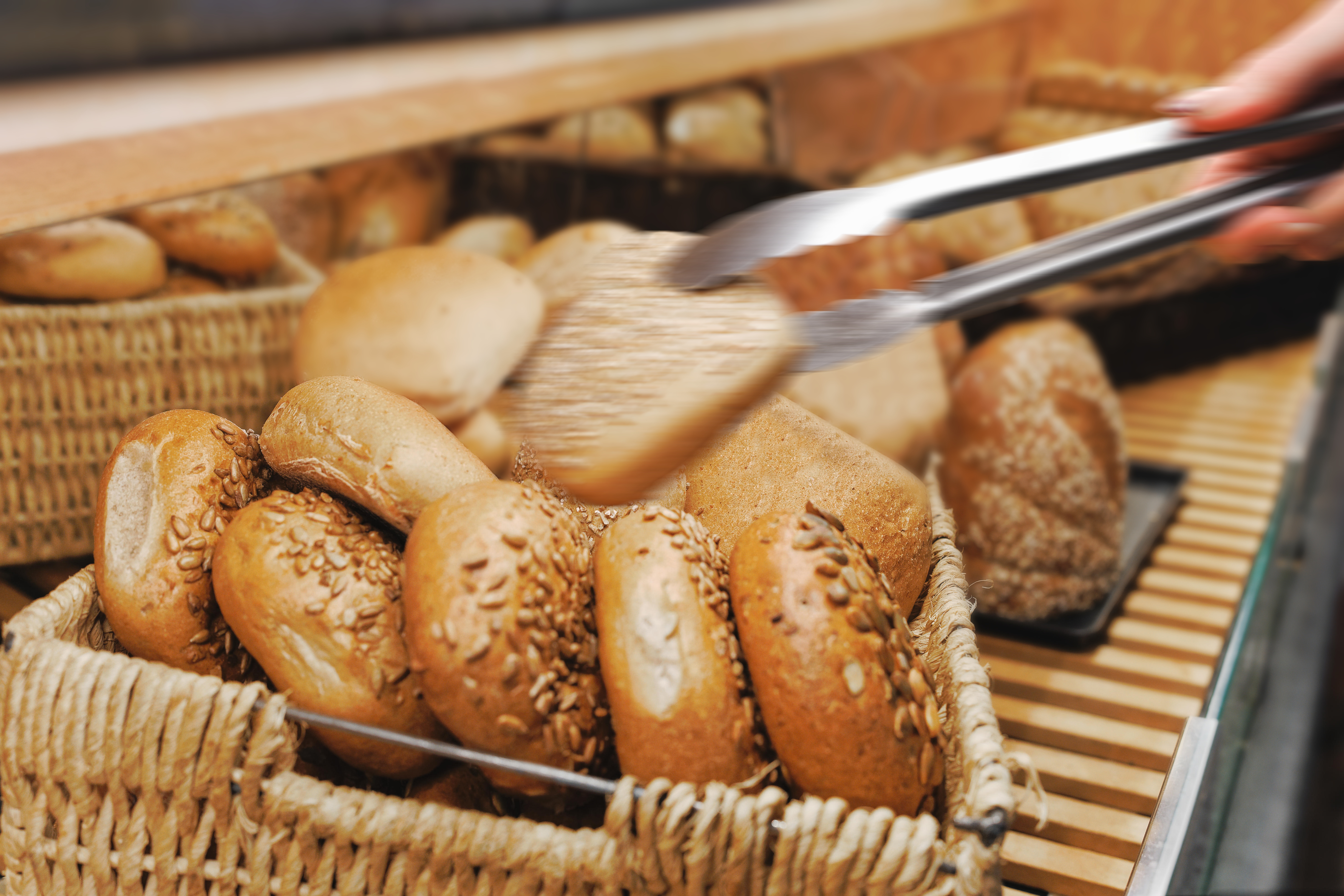 Hand using tongs to pick a seeded bread roll from a basket filled with various fresh bread rolls.