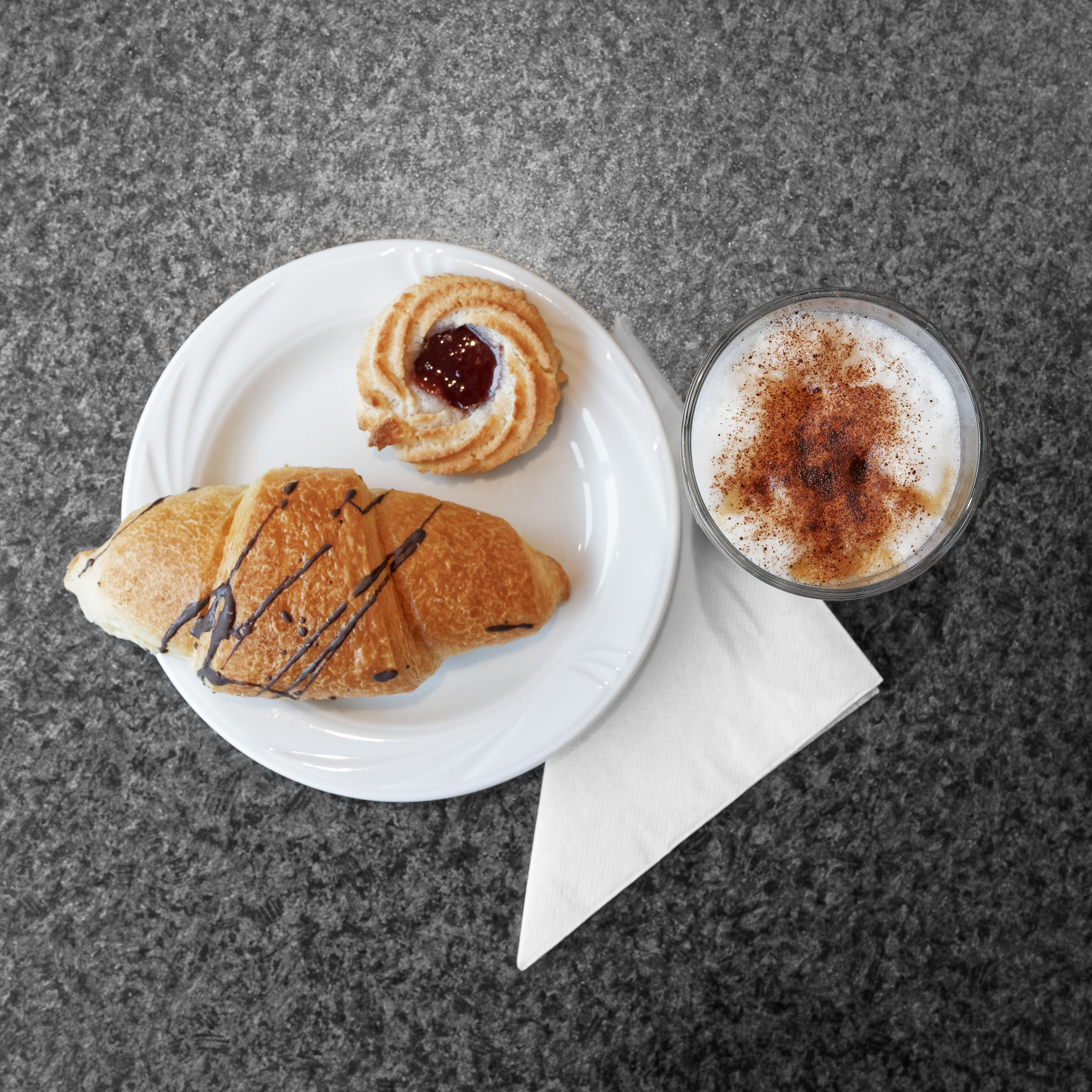 White plate with a croissant drizzled with chocolate and a cookie topped with jam next to a glass of frothy coffee with cinnamon on a gray countertop.