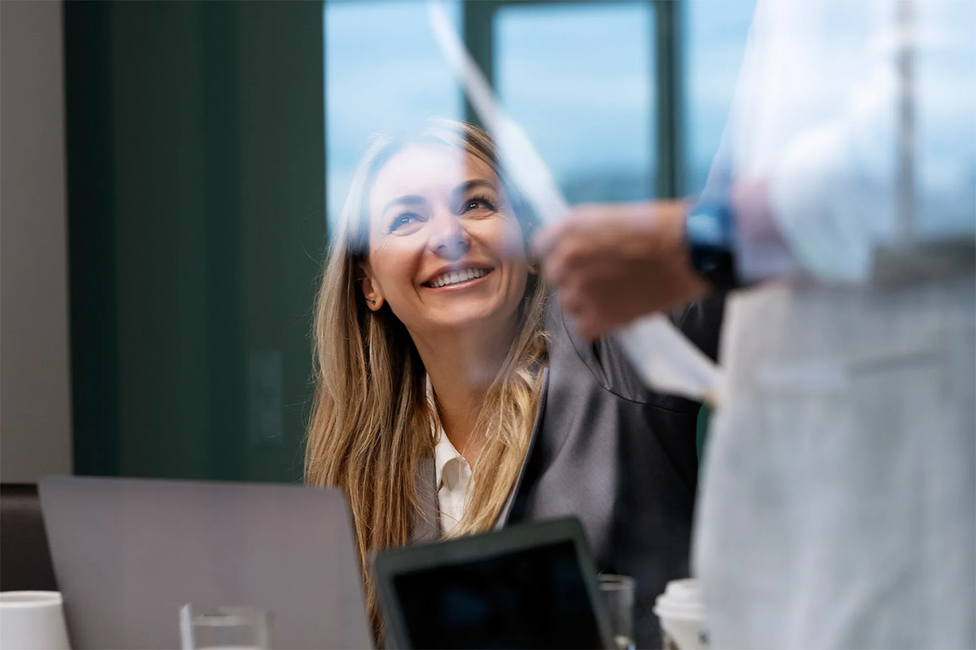 Smiling woman in a business suit looks up while sitting at a table with laptops in an office.