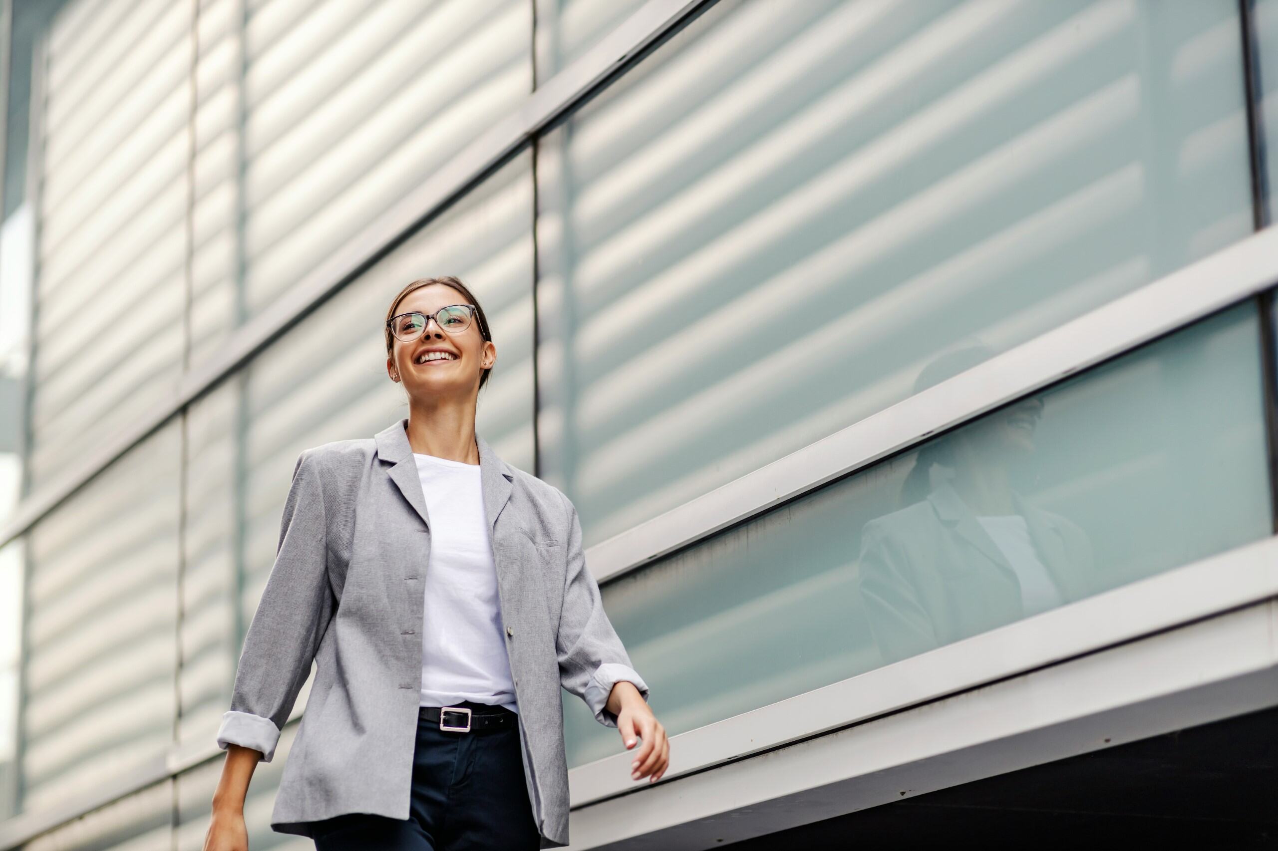 Smiling young woman in glasses wearing grey blazer and white shirt walking by modern glass building.