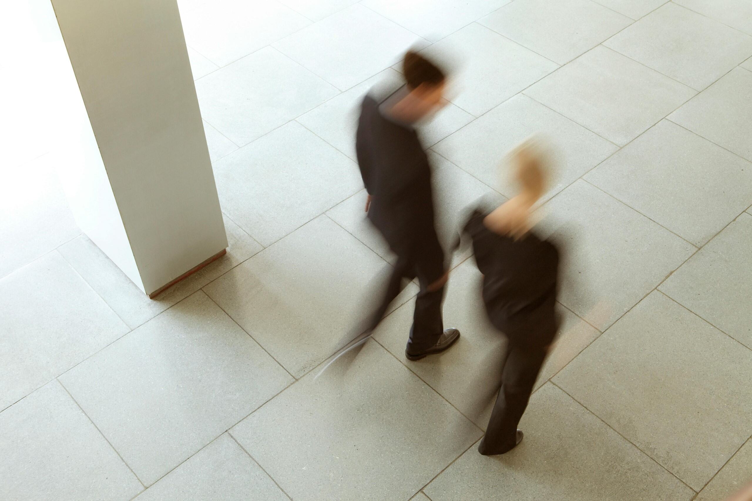 Blurred motion of two people walking past a large white pillar on a tiled floor.