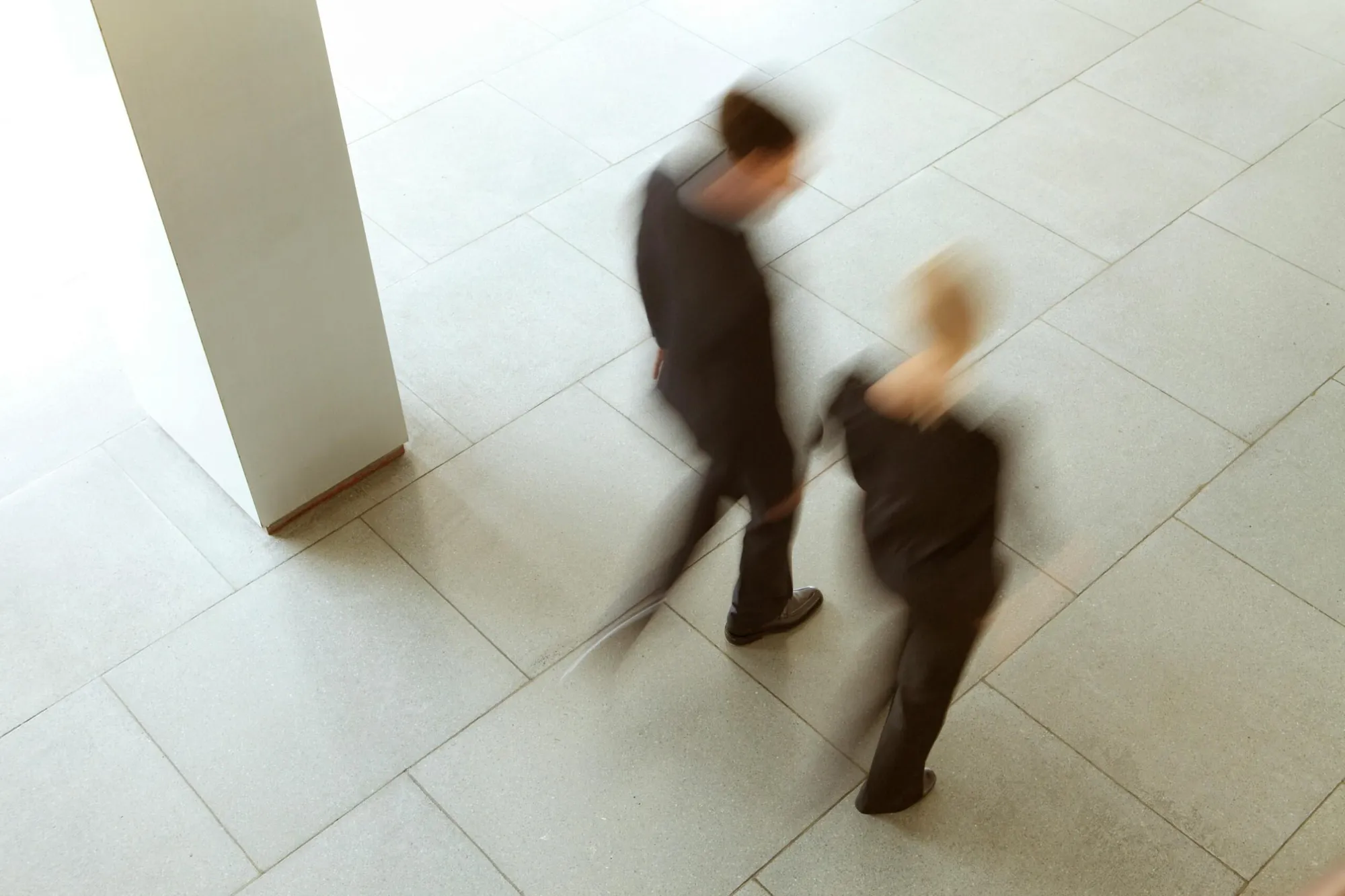 Zwei Personen gehen in Bewegungsunschärfe an einer großen weißen Säule über gefliesten Boden vorbei