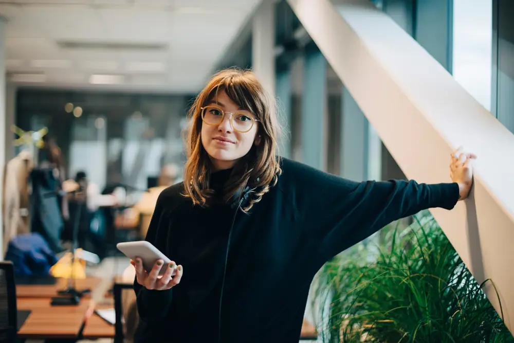 Young woman with glasses holding a smartphone, leaning on a window sill in an office setting.