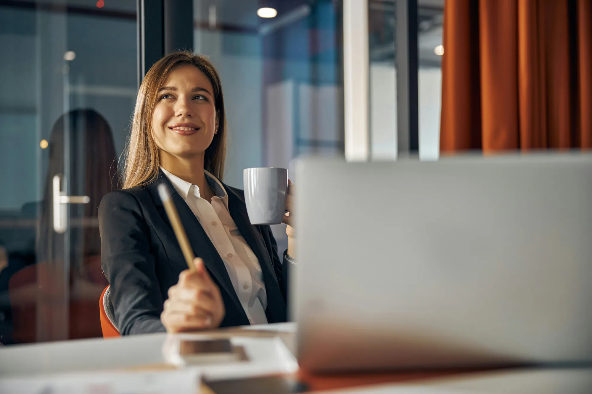 Lächelnde Geschäftsfrau mit Tasse und Stift am Schreibtisch mit Laptop im Büro
