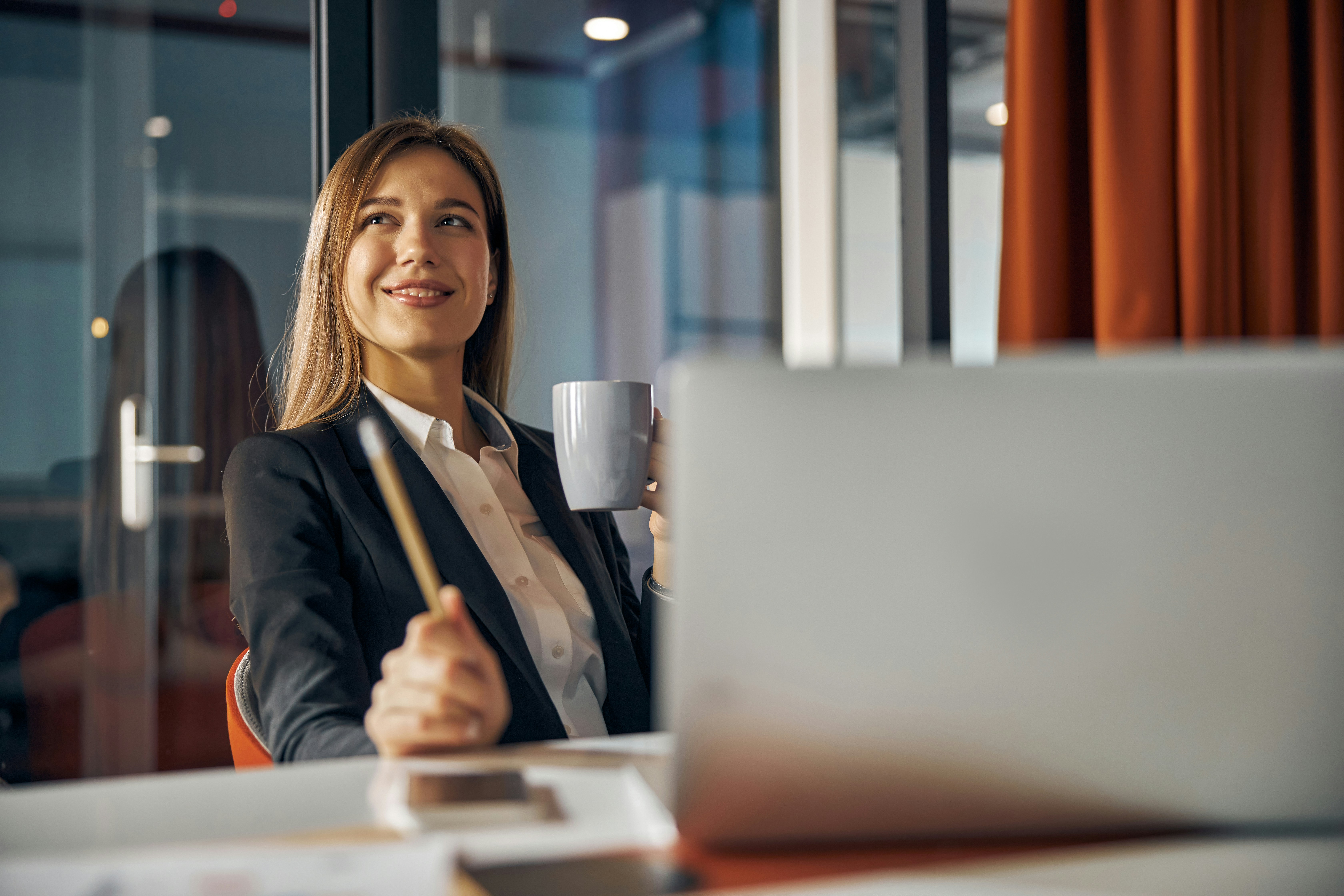 Smiling businesswoman holding a mug and pencil seated at a desk with a laptop in an office.