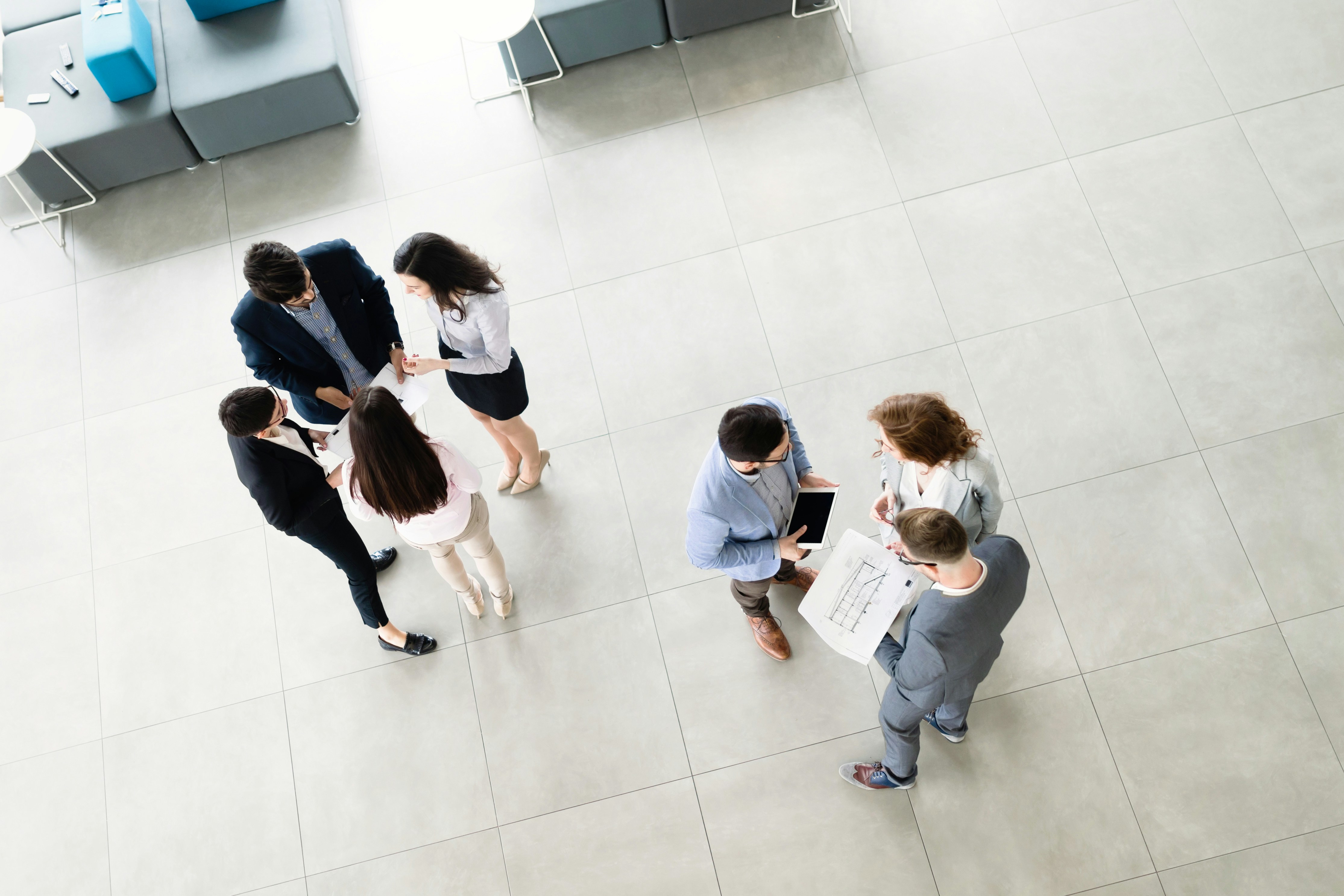 Two small groups of business people stand in an office lobby discussing documents and a tablet.