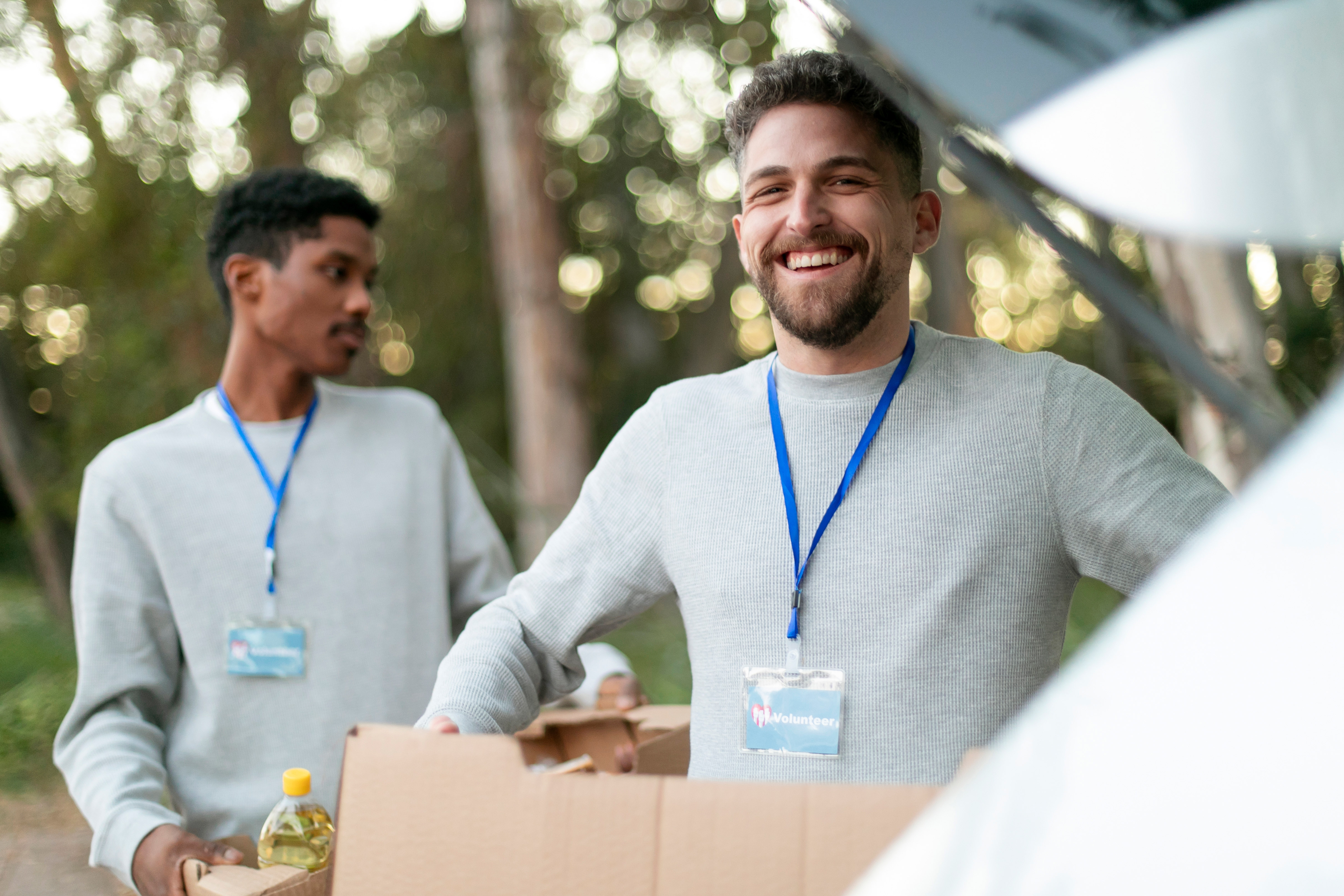 Two male volunteers wearing grey sweaters and blue badges carry boxes outdoors in a wooded area.