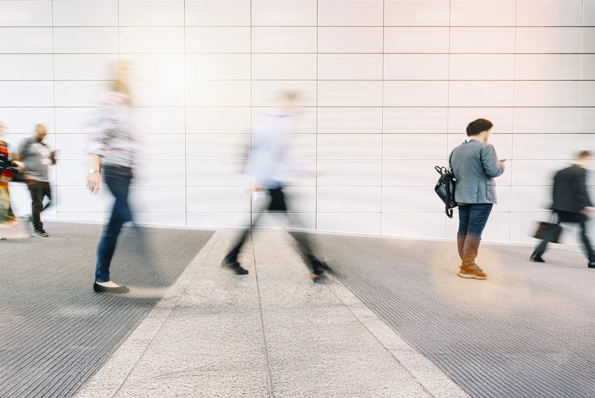 Blurred figures walking in a modern indoor space with one person standing and looking at a phone.