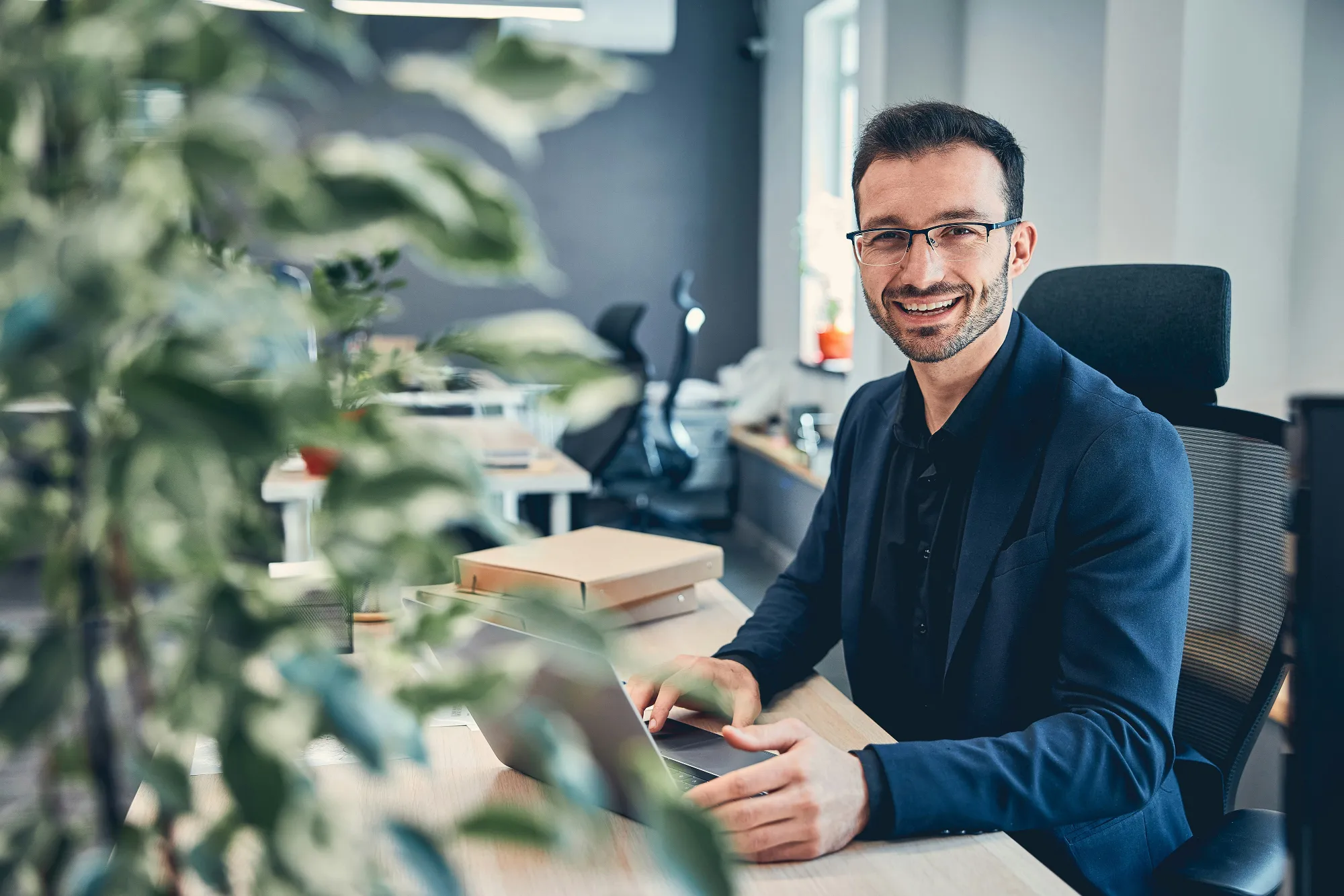 Lächelnder Mann mit Brille und Sakko arbeitet am Laptop in einem modernen Büro