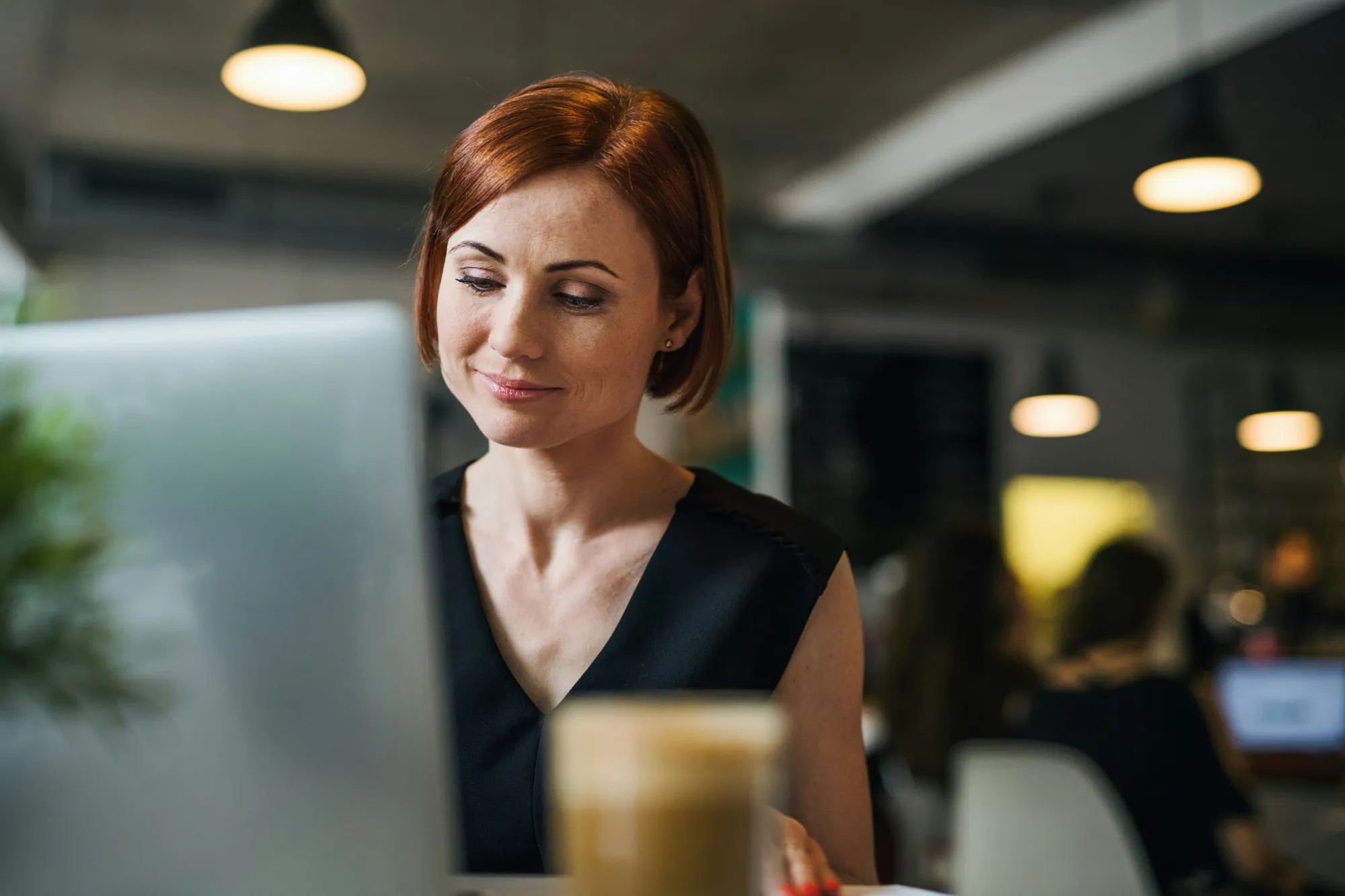 Frau mit kurzen roten Haaren arbeitet im Café am Laptop, davor eine Tasse Kaffee