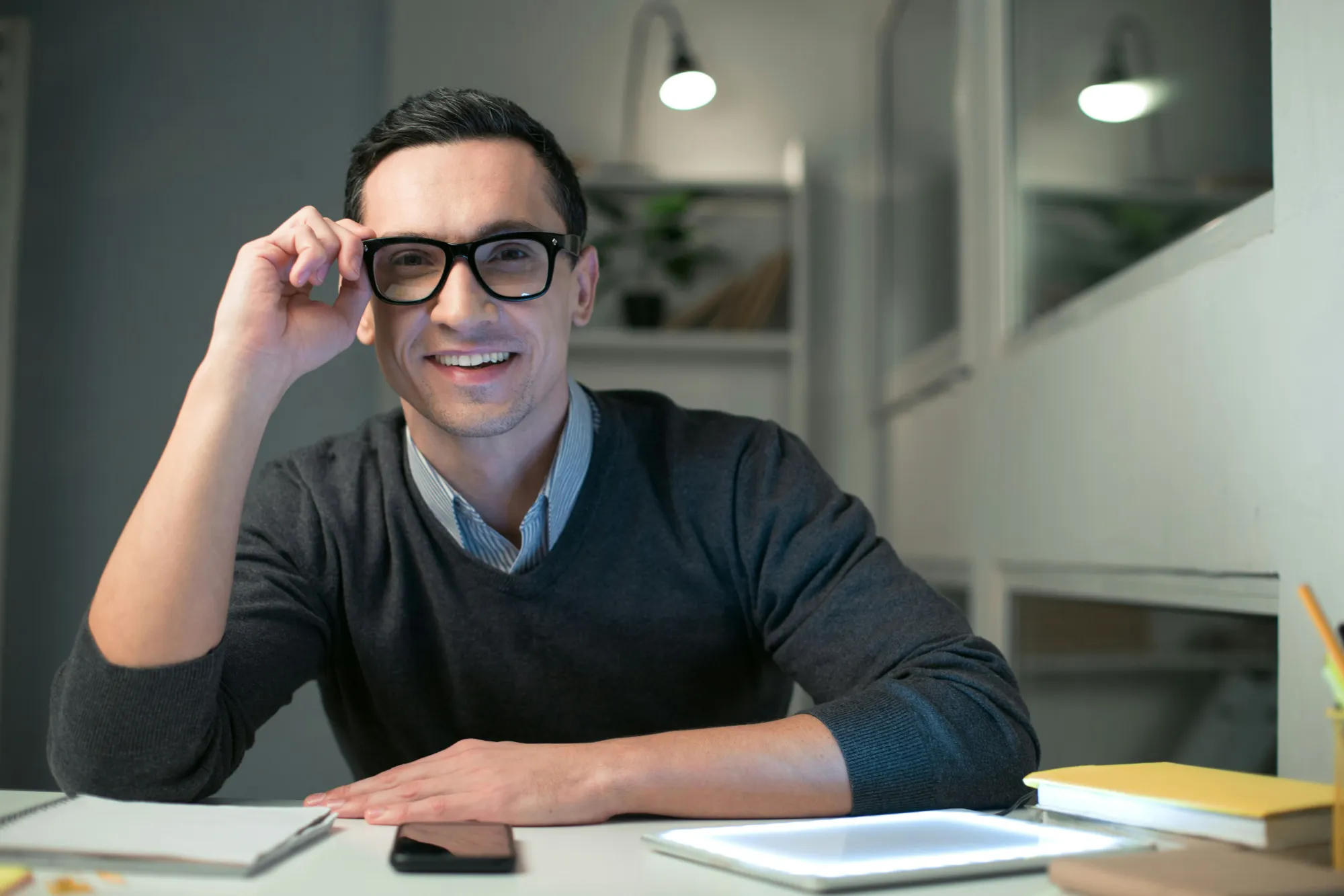 Lächelnder Mann mit Brille am Schreibtisch mit Tablet, Smartphone y Notizbüchern in modernem Raum