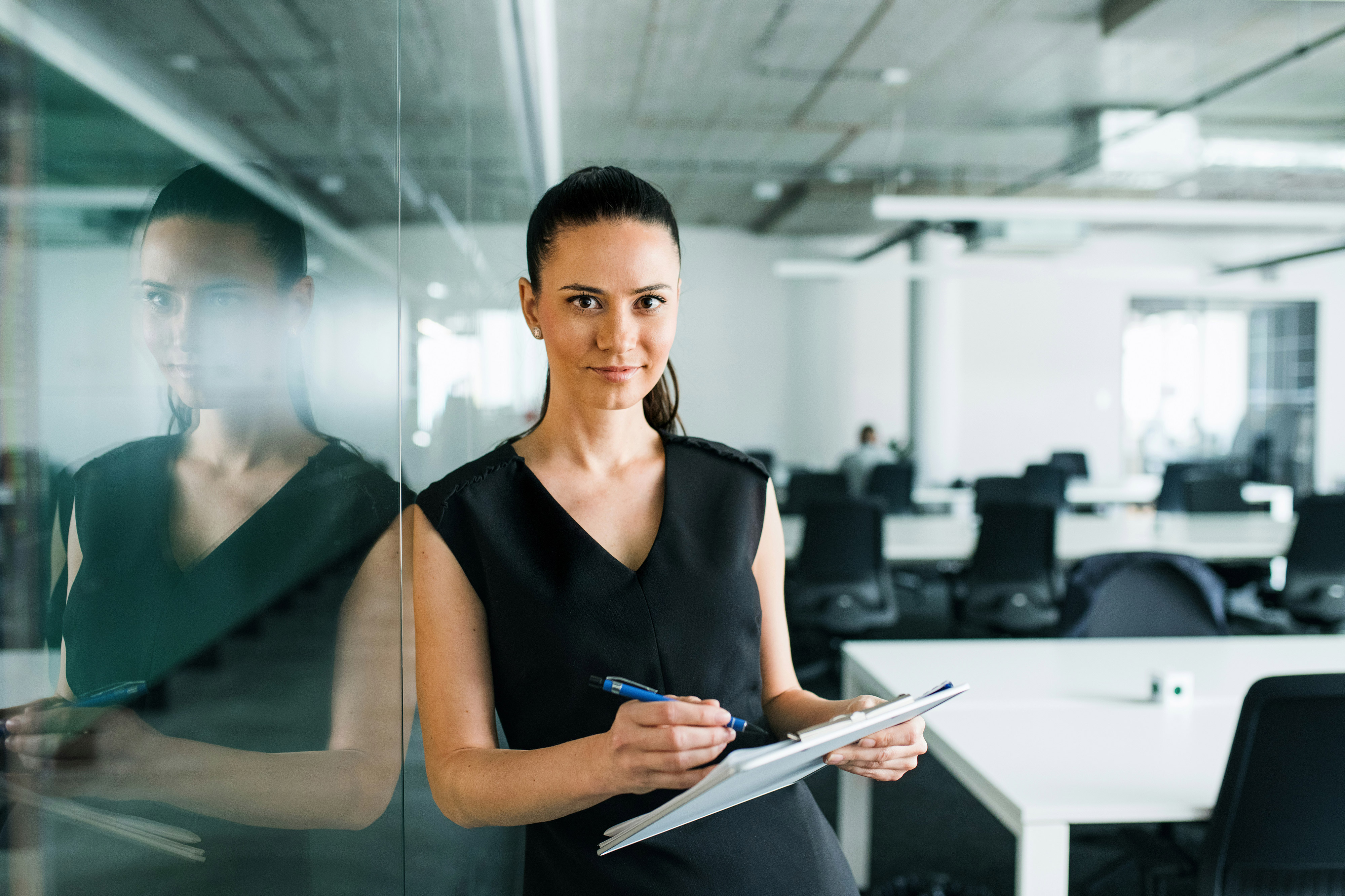 Woman holding a clipboard and pen standing by a glass wall in a modern office.