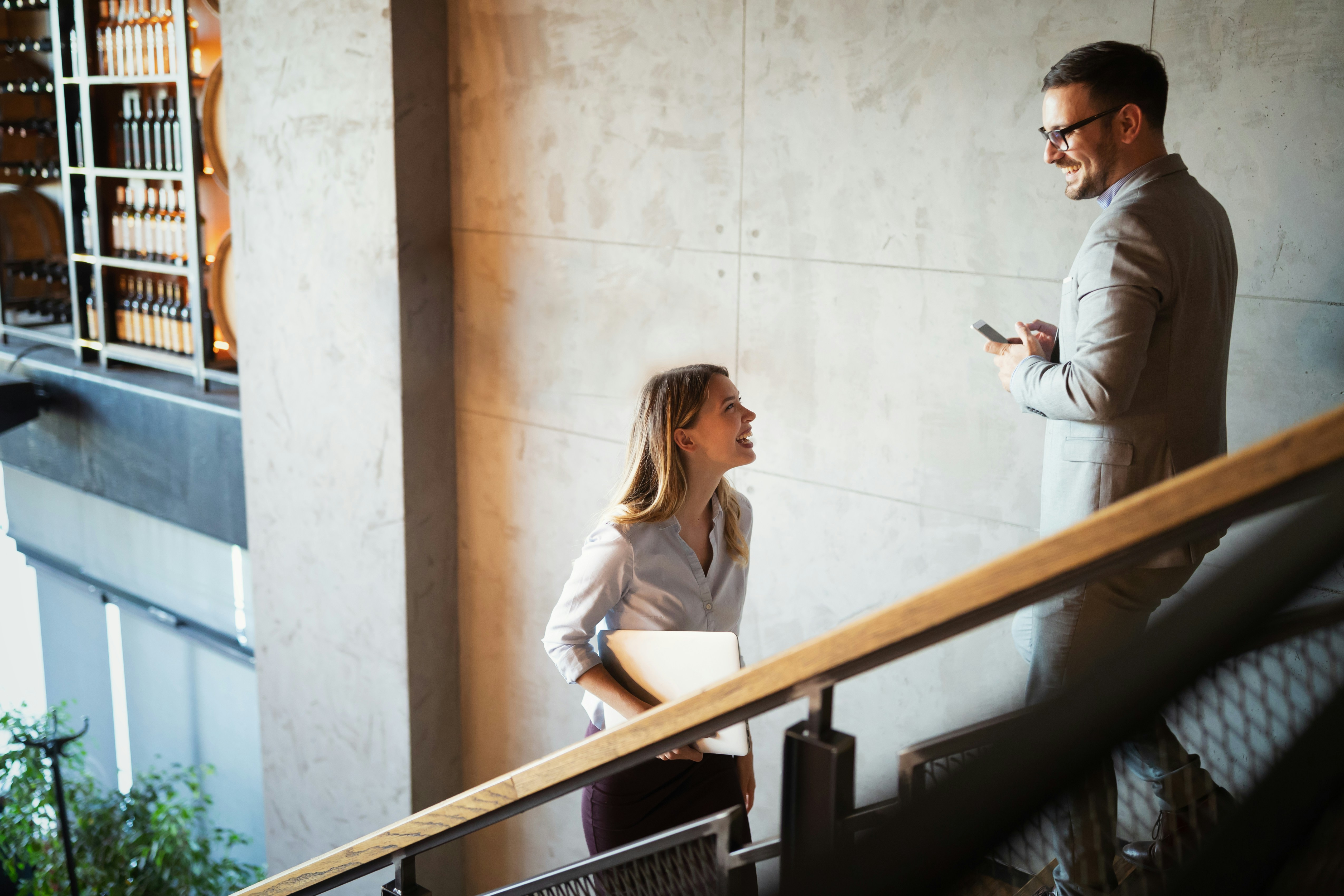 Woman holding laptop talks and smiles with man in suit standing on stairs in modern building.