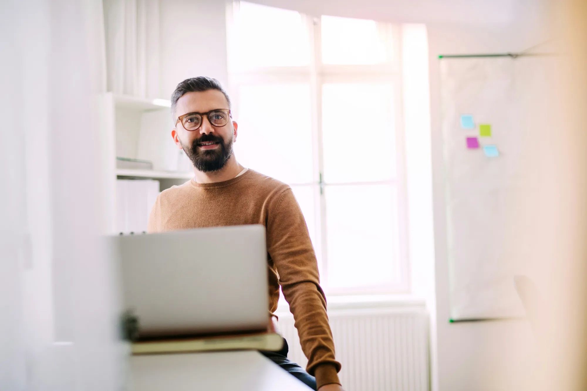 Mann mit Brille et braunem Pullover am Schreibtisch mit Laptop im hellen Büro