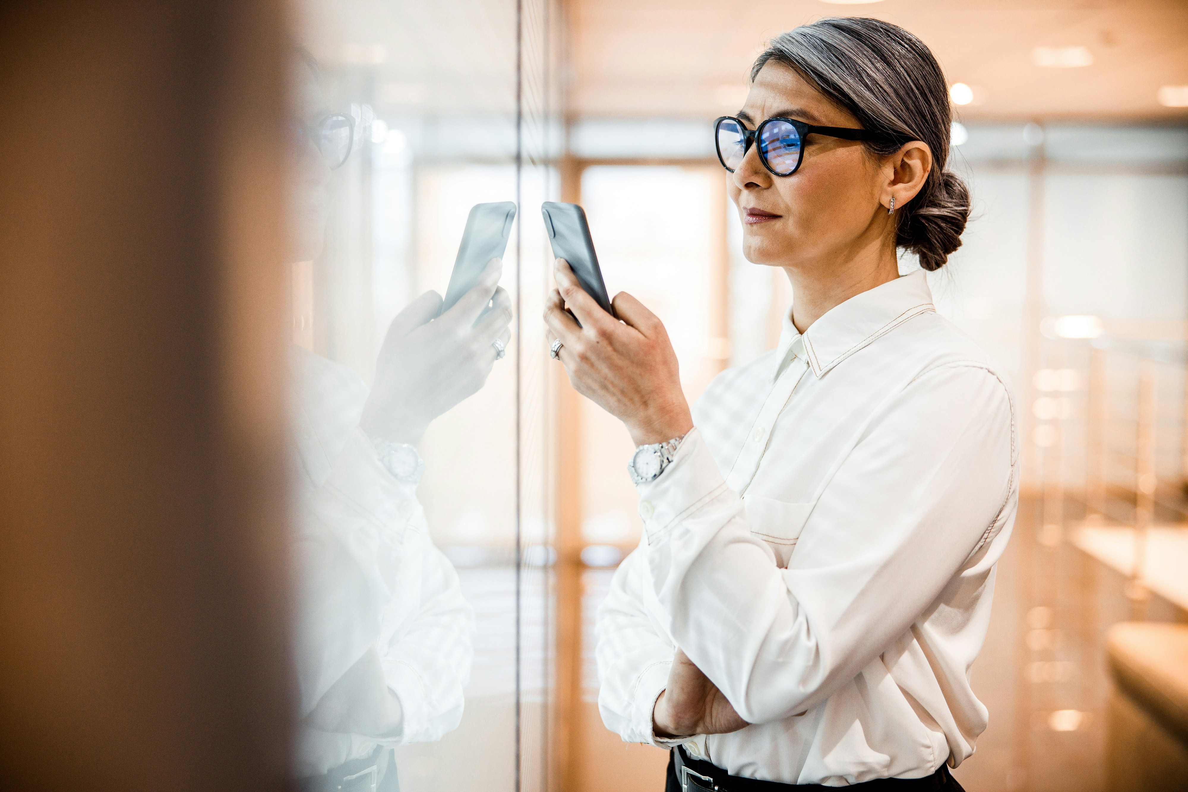 Woman in glasses and white shirt looking at her phone next to a reflective glass wall.