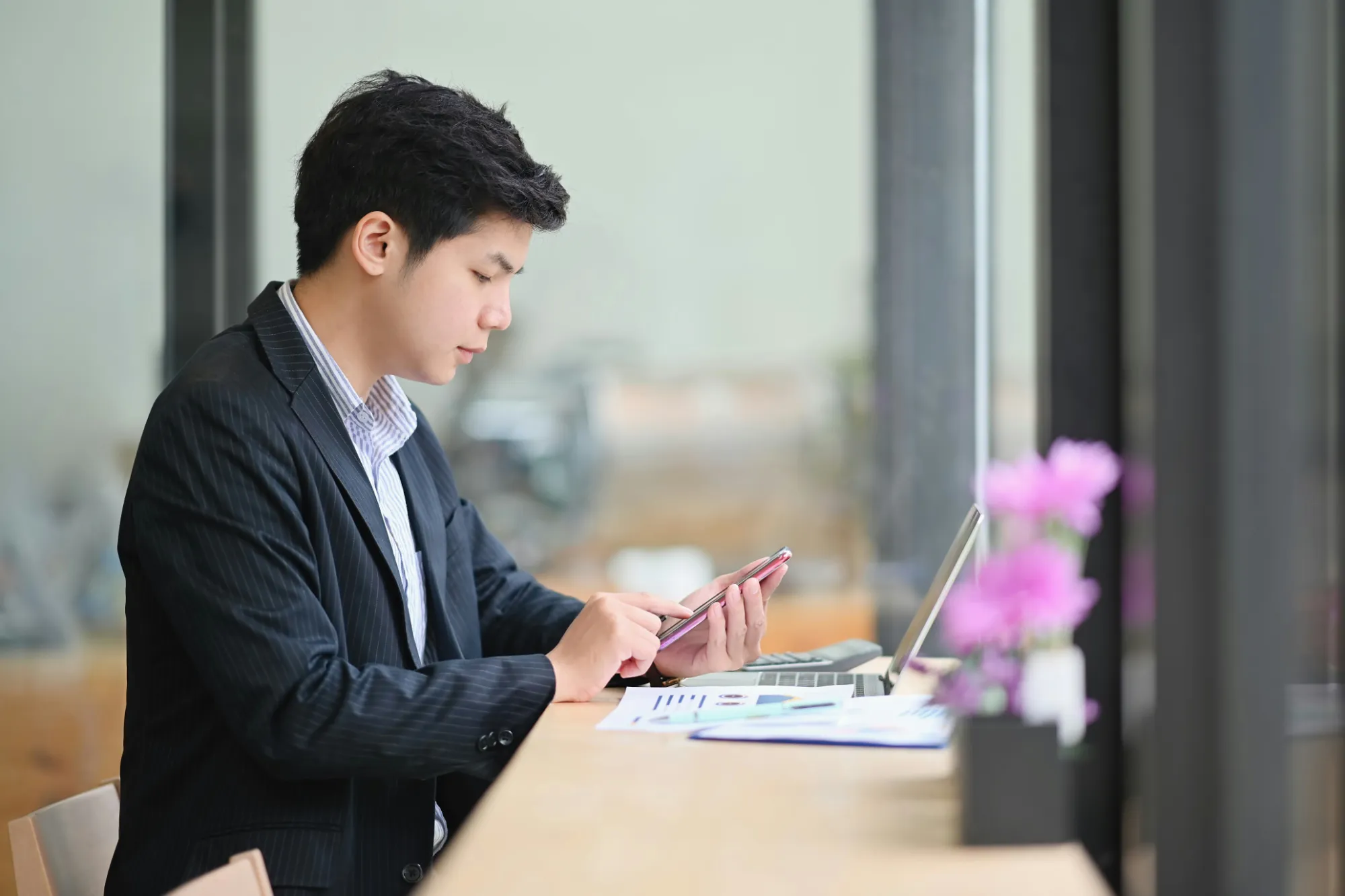 Jovem de terno usando um smartphone enquanto está sentado em uma mesa com um laptop e papéis.