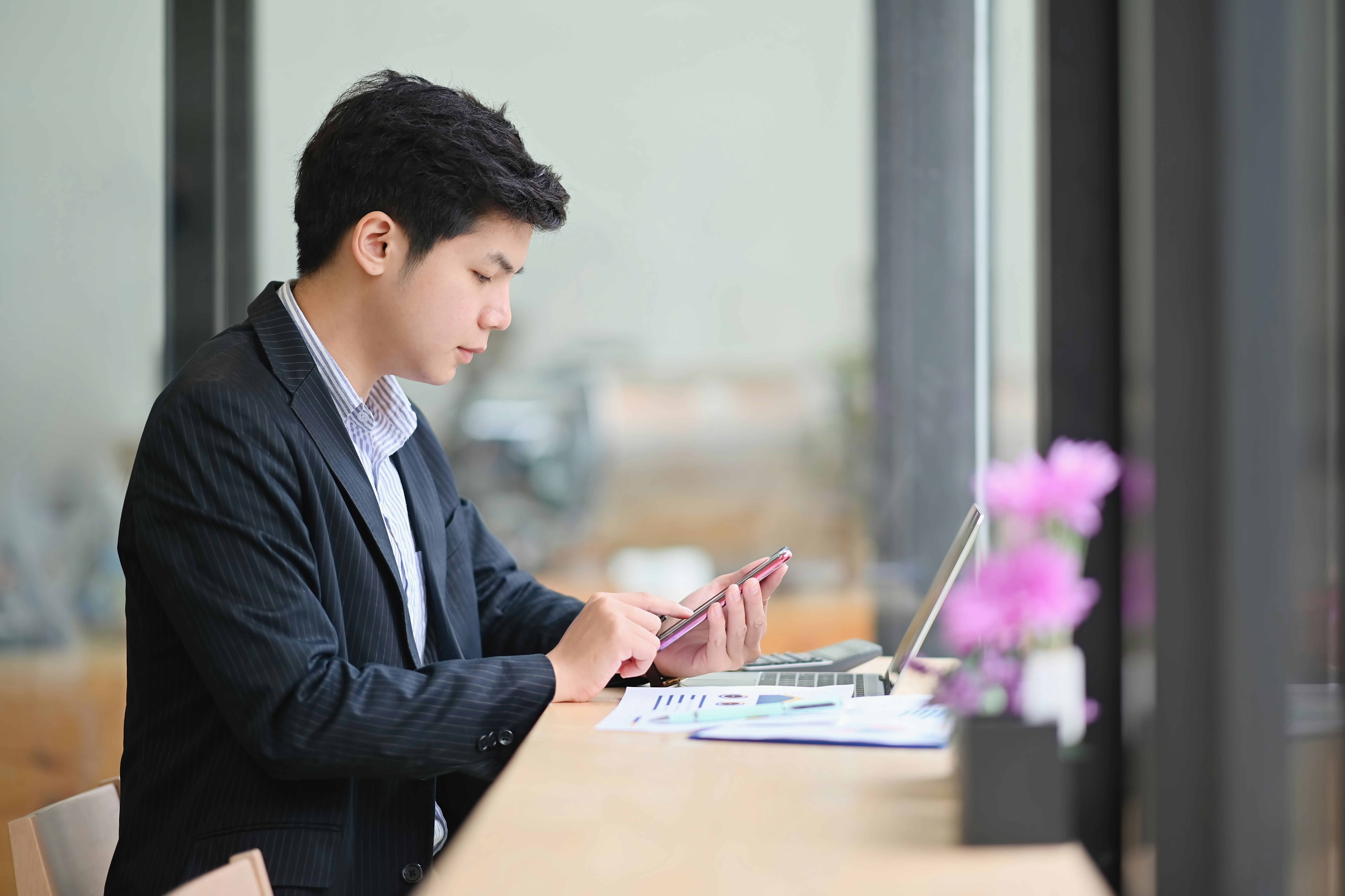 Young man in a suit using a smartphone while sitting at a desk with a laptop and papers.