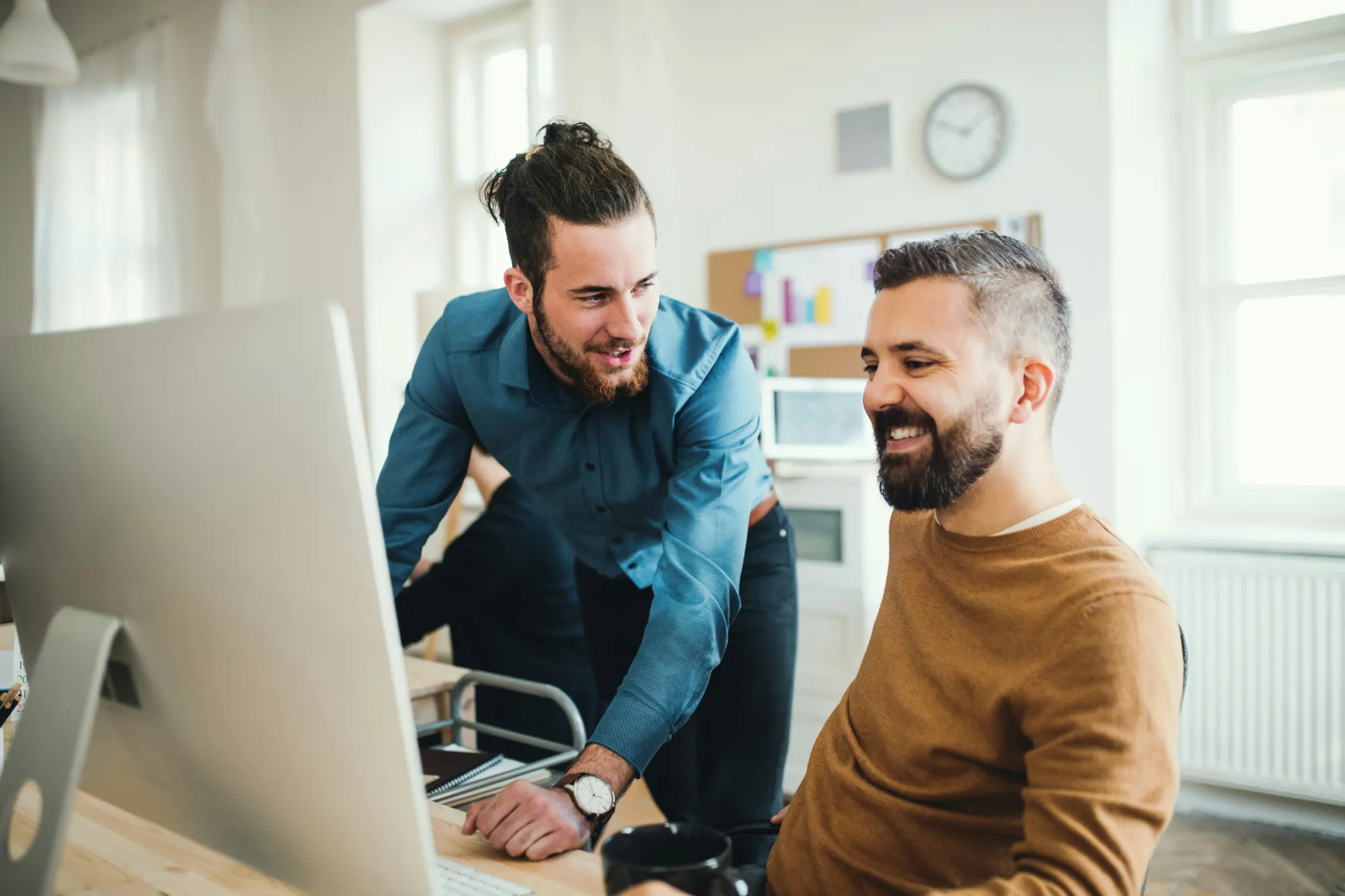 Dos hombres arbeiten lächelnd zusammen am Desktop-Computer in einem hellen Büro.