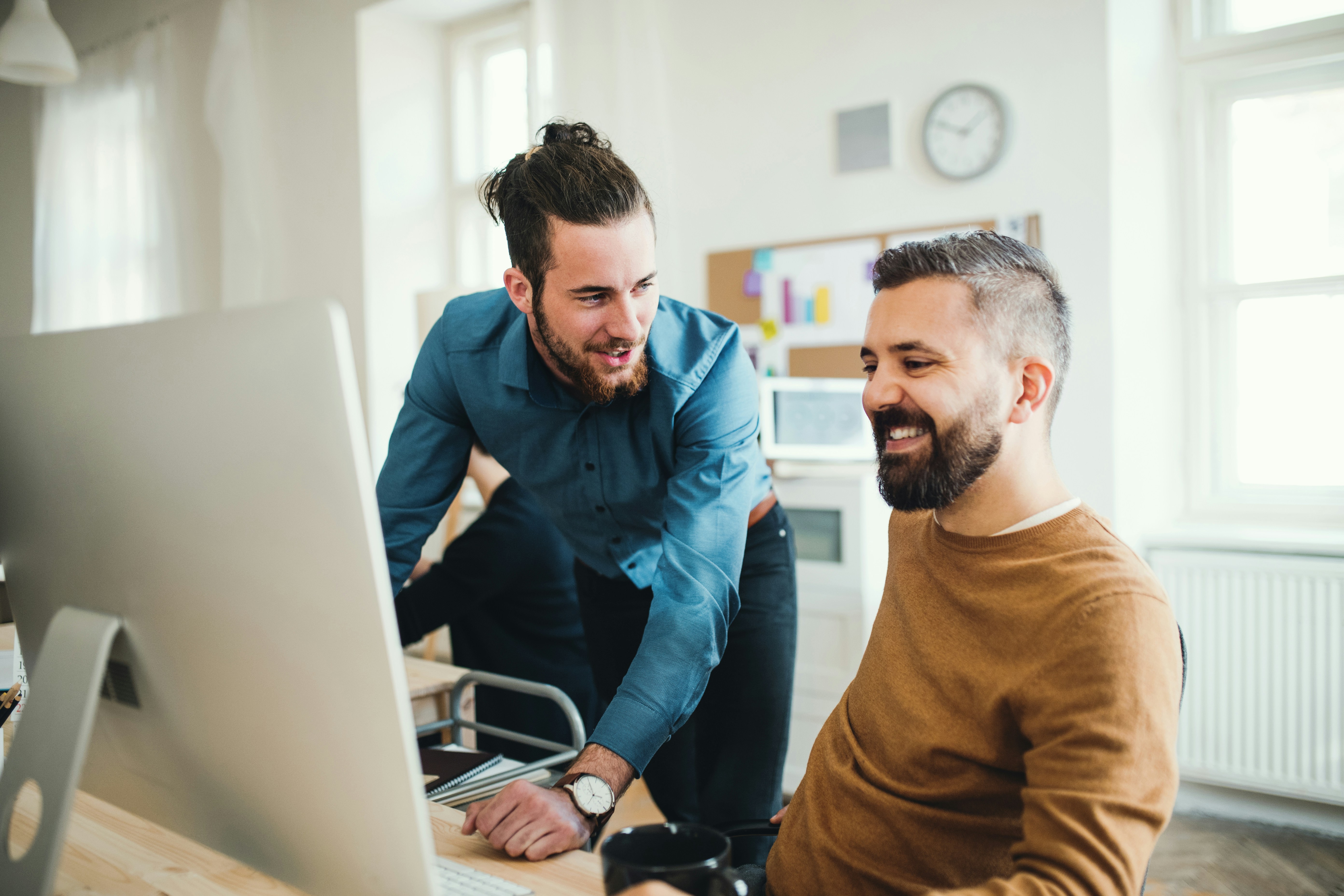 Two men collaborating and smiling while working at a desktop computer in a bright office.