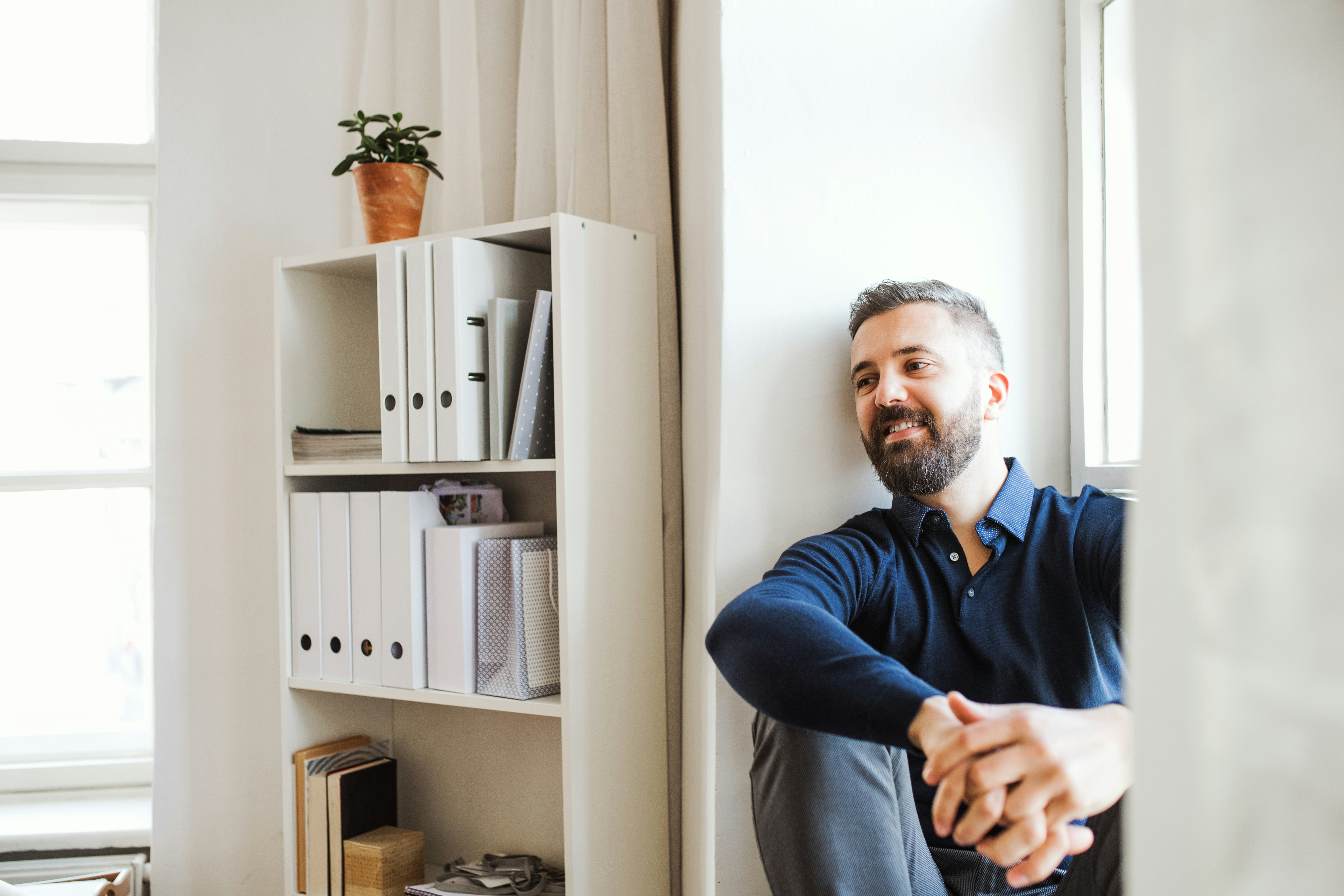 Smiling bearded man sitting by a window in a bright room with a white shelf and plant.